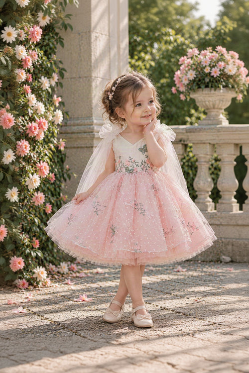 A young girl stands outdoors in a garden wearing a light pink tulle flower girl dress with a white bodice, floral embroidery, and a sheer white veil. She is posed in front of a stone balustrade and a flower-covered wall.