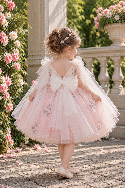 A young girl with her hair in a bun stands outdoors in a garden wearing a light pink tulle flower girl dress with a large white bow on the back, sheer flutter sleeves, and floral embroidery. She is facing away from the camera, looking to the side.