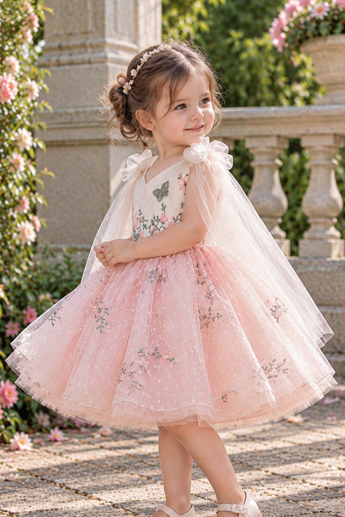 A young girl wearing a pink and white tulle flower girl dress with butterfly and floral embroidery, standing in a garden courtyard.