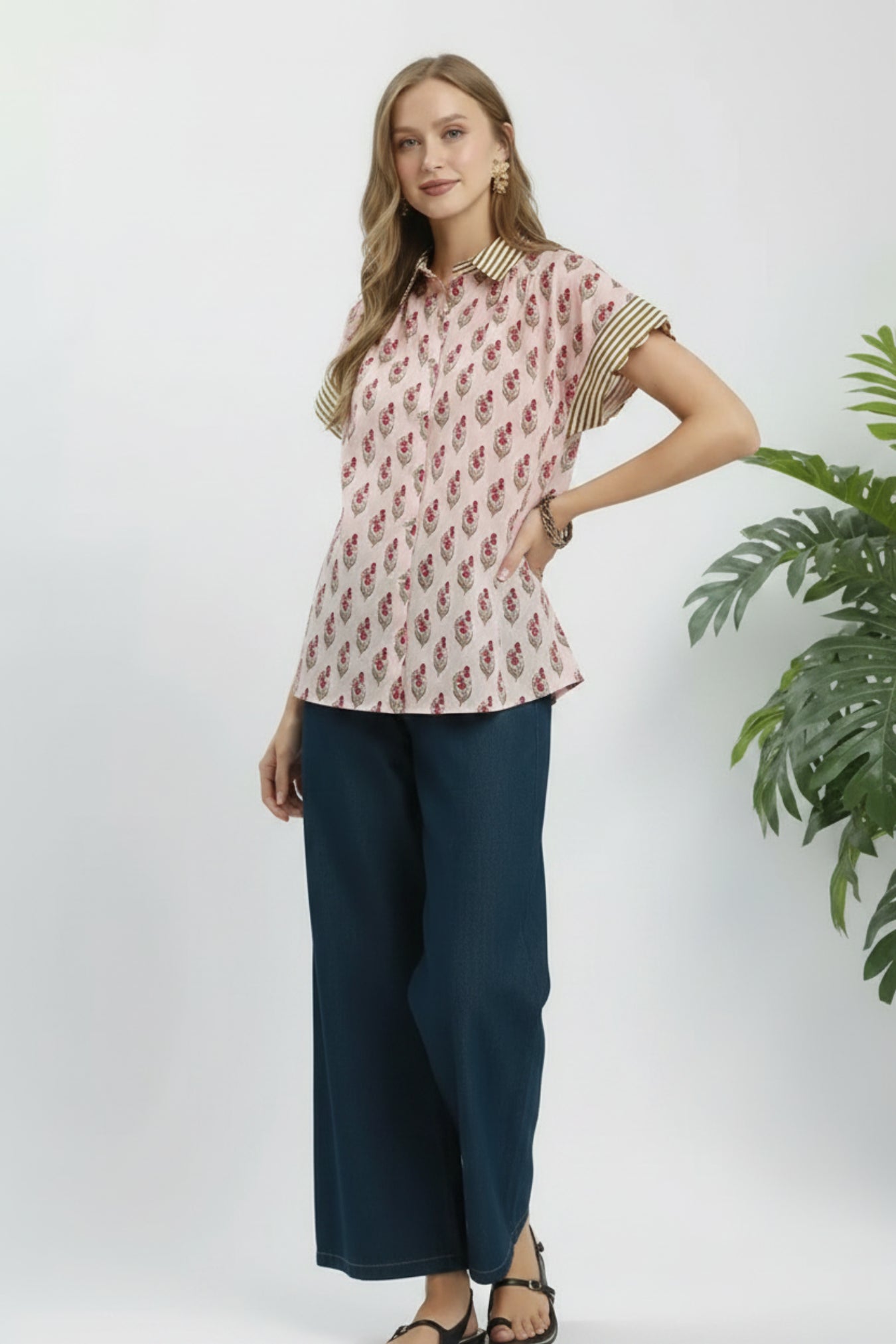 Woman wearing a patterned top and dark pants on a white background next to a plant