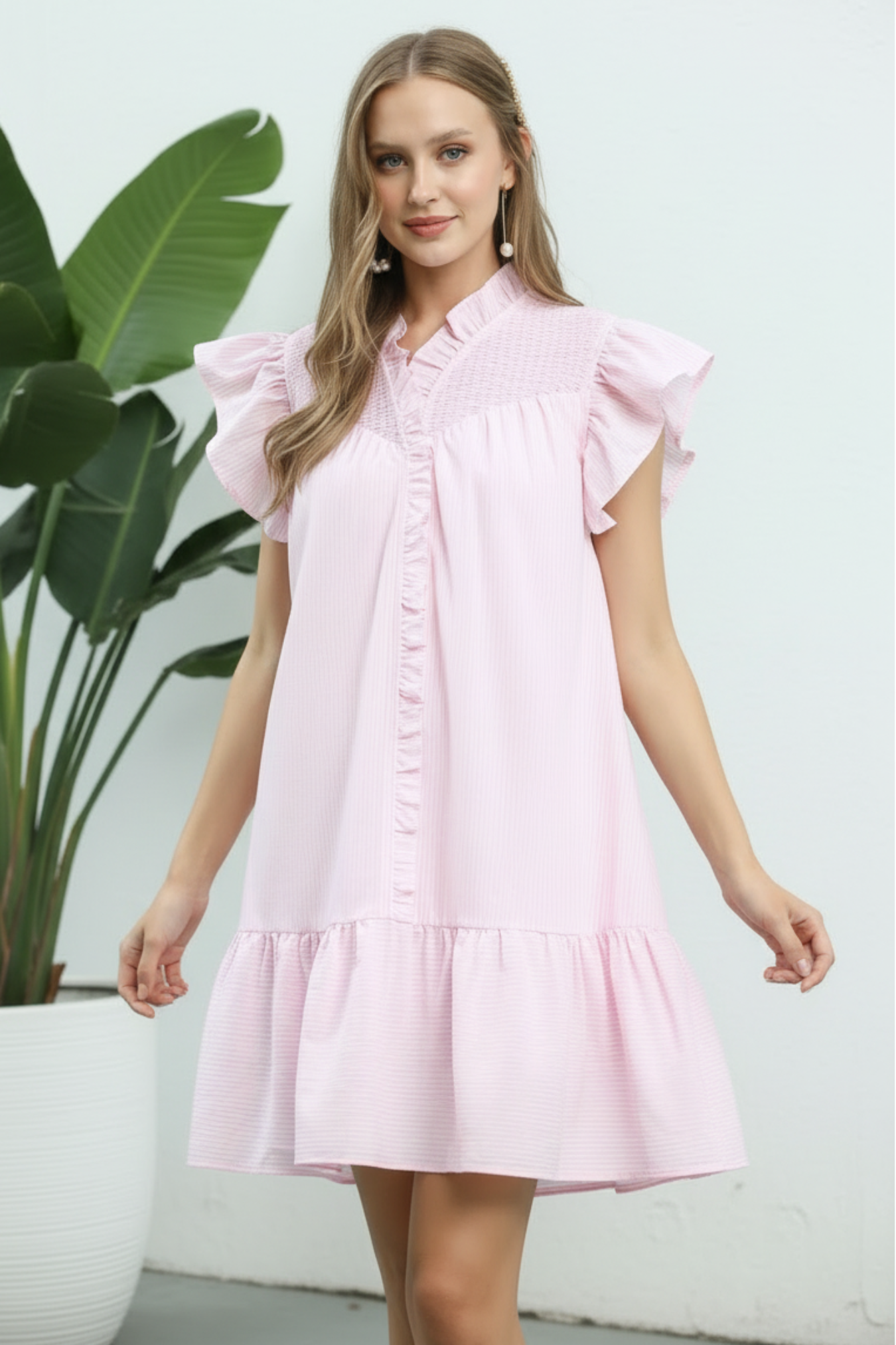 Woman wearing a pink dress with ruffled sleeves against a white background next to a plant