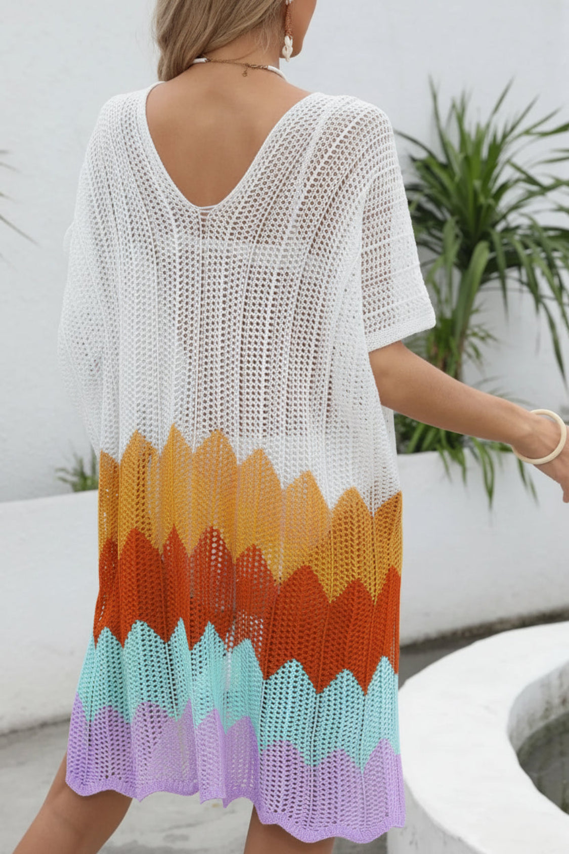 Woman wearing a colorful crochet cover-up with a plant and white wall in the background