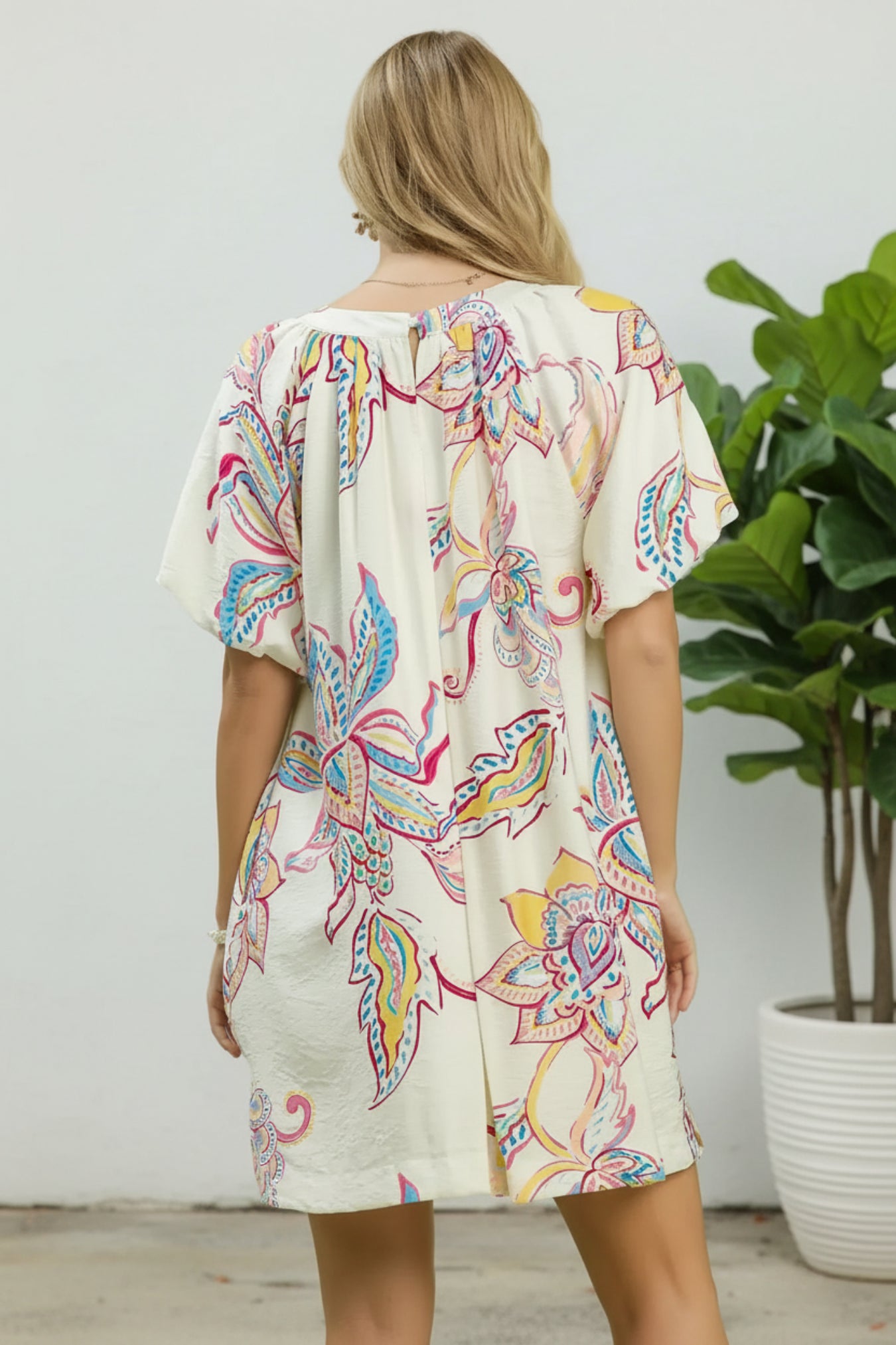 Woman wearing a colorful patterned dress standing in a room with a plant.