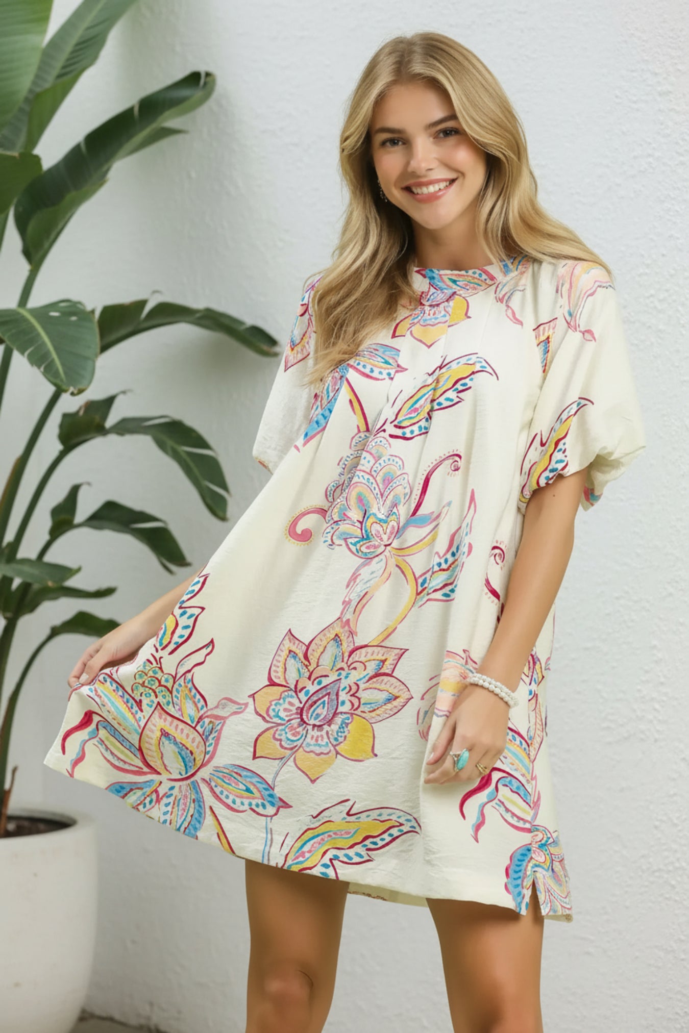 Woman wearing a colorful floral dress standing next to a potted plant against a white wall.