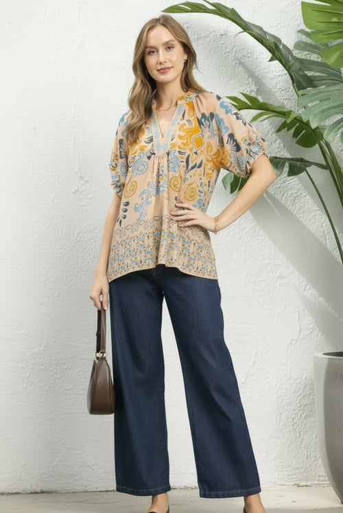Woman wearing a patterned blouse and navy pants against a plant and white background