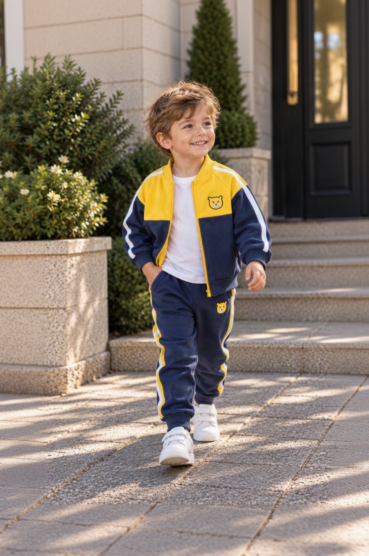 A young boy walking outdoors on stone steps wearing a navy and yellow athletic tracksuit with a white t-shirt and white sneakers.