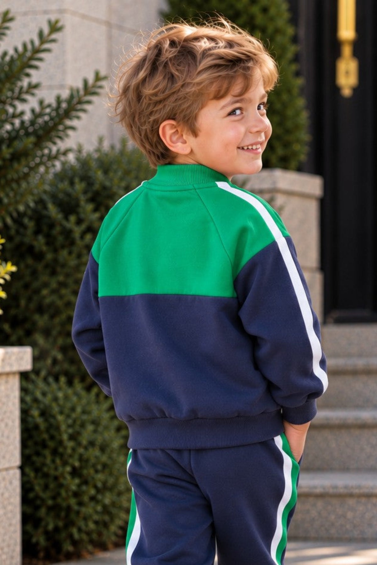 A young boy wearing a green, navy, and white two-piece tracksuit, standing on stone steps in front of a building.