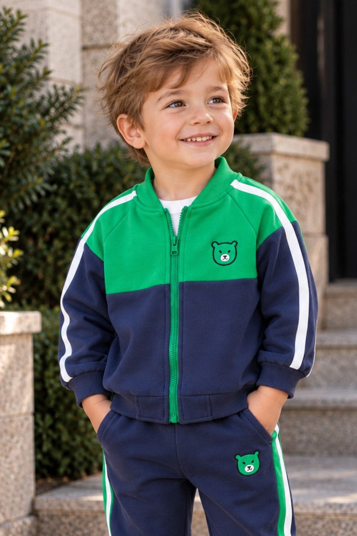 A young boy wearing a two-piece navy blue and green tracksuit with white stripes, standing outdoors on stone steps in front of a building.