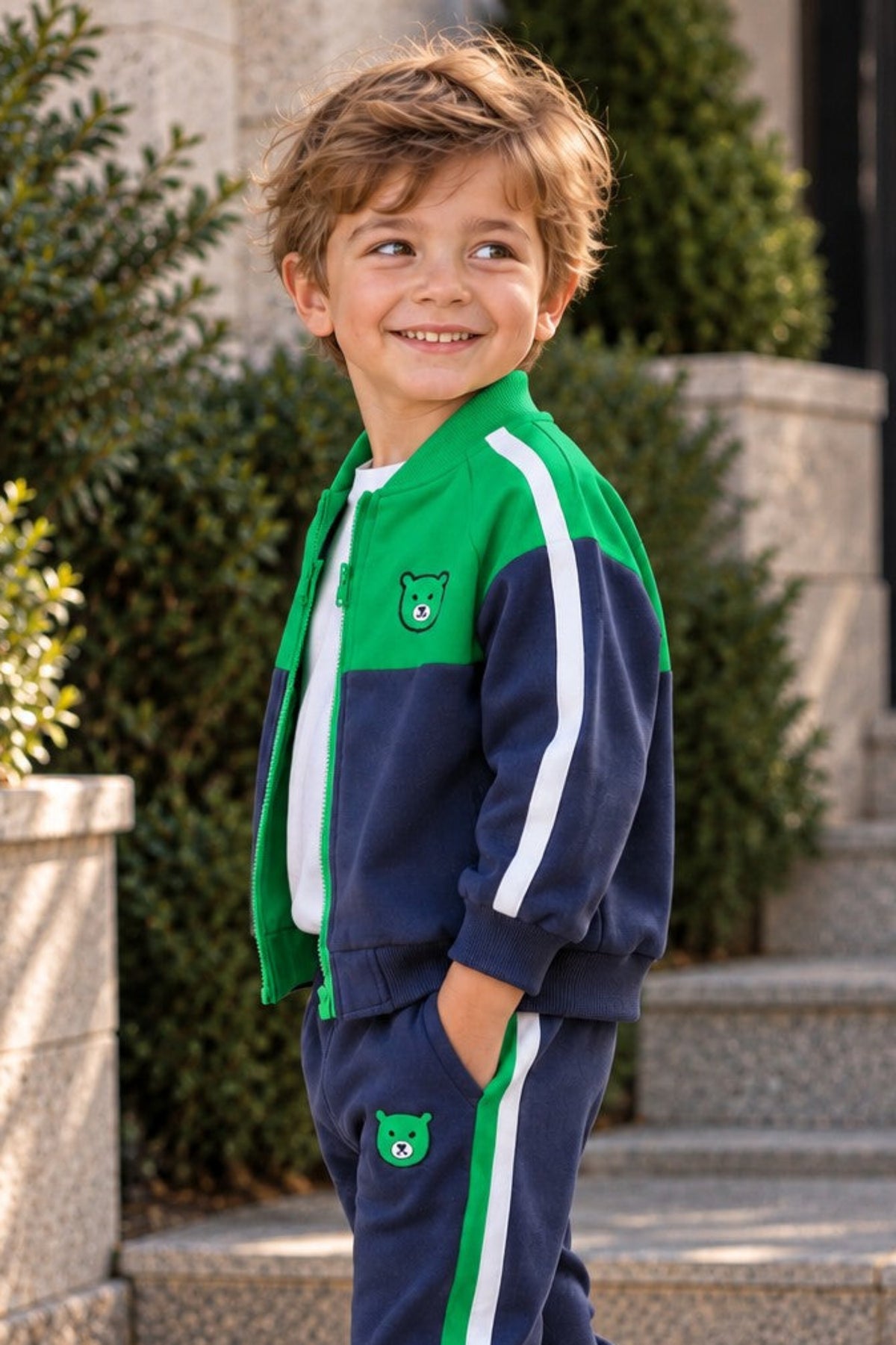 A young boy wearing a navy blue and green tracksuit with white stripes, standing on stone steps outdoors.