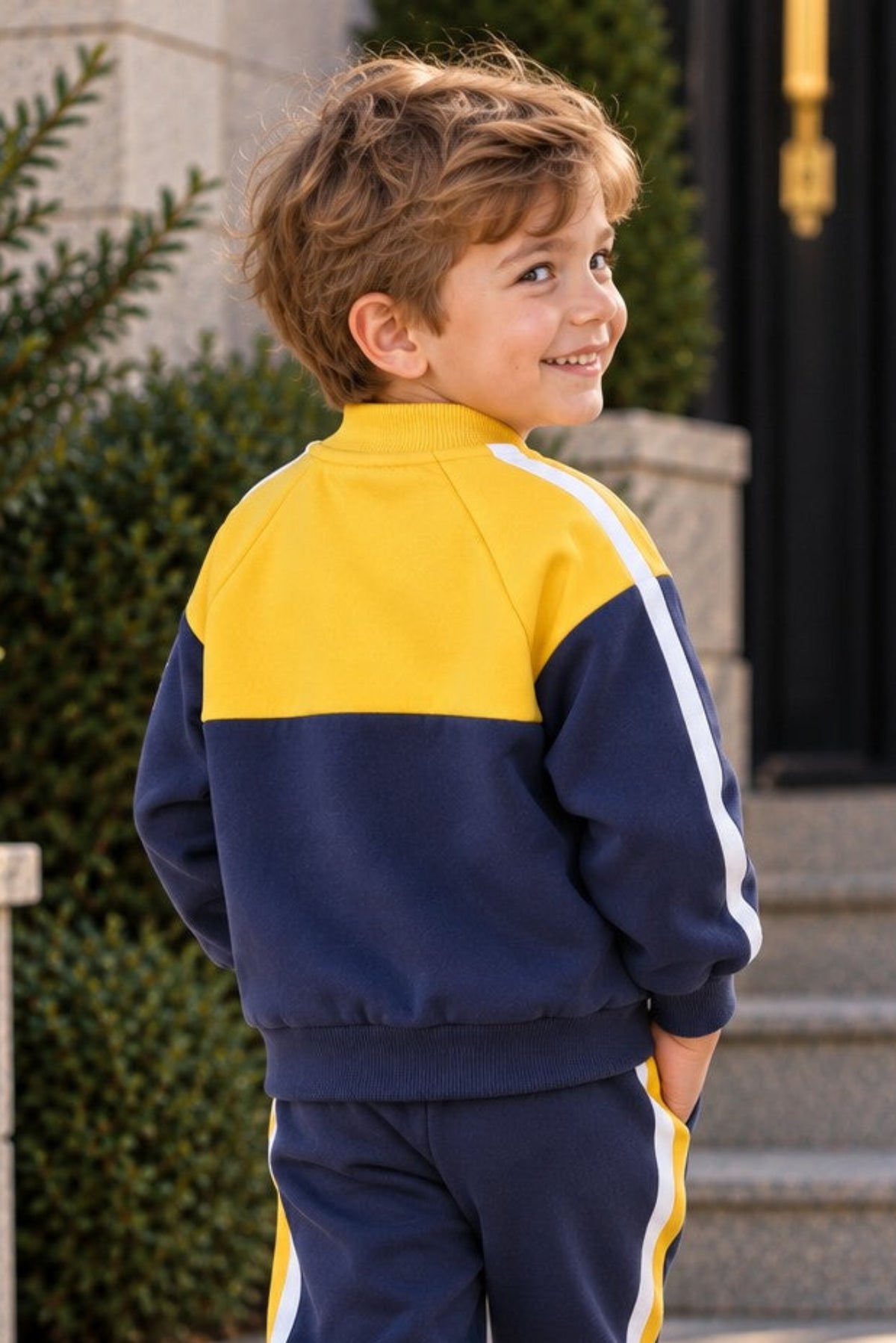 A young boy wearing a navy blue and yellow color-blocked tracksuit with white side stripes, standing on stone steps outdoors.