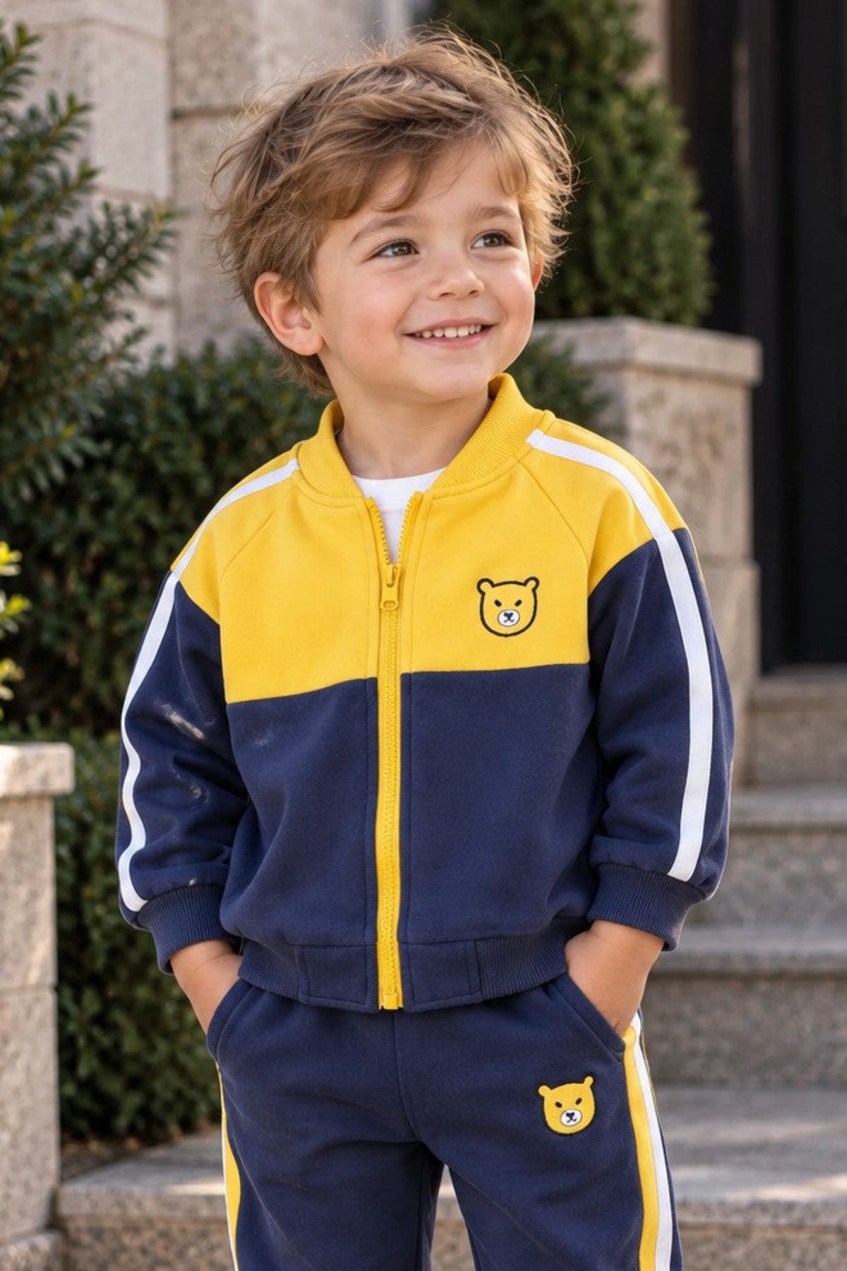 A young boy wearing a navy blue and yellow color-blocked tracksuit with white sneakers, standing outdoors on stone steps in front of a building.