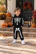 A young boy wearing a black skeleton-themed hoodie and sweatpants set, standing on stone steps in front of a house decorated with pumpkins and autumn leaves.