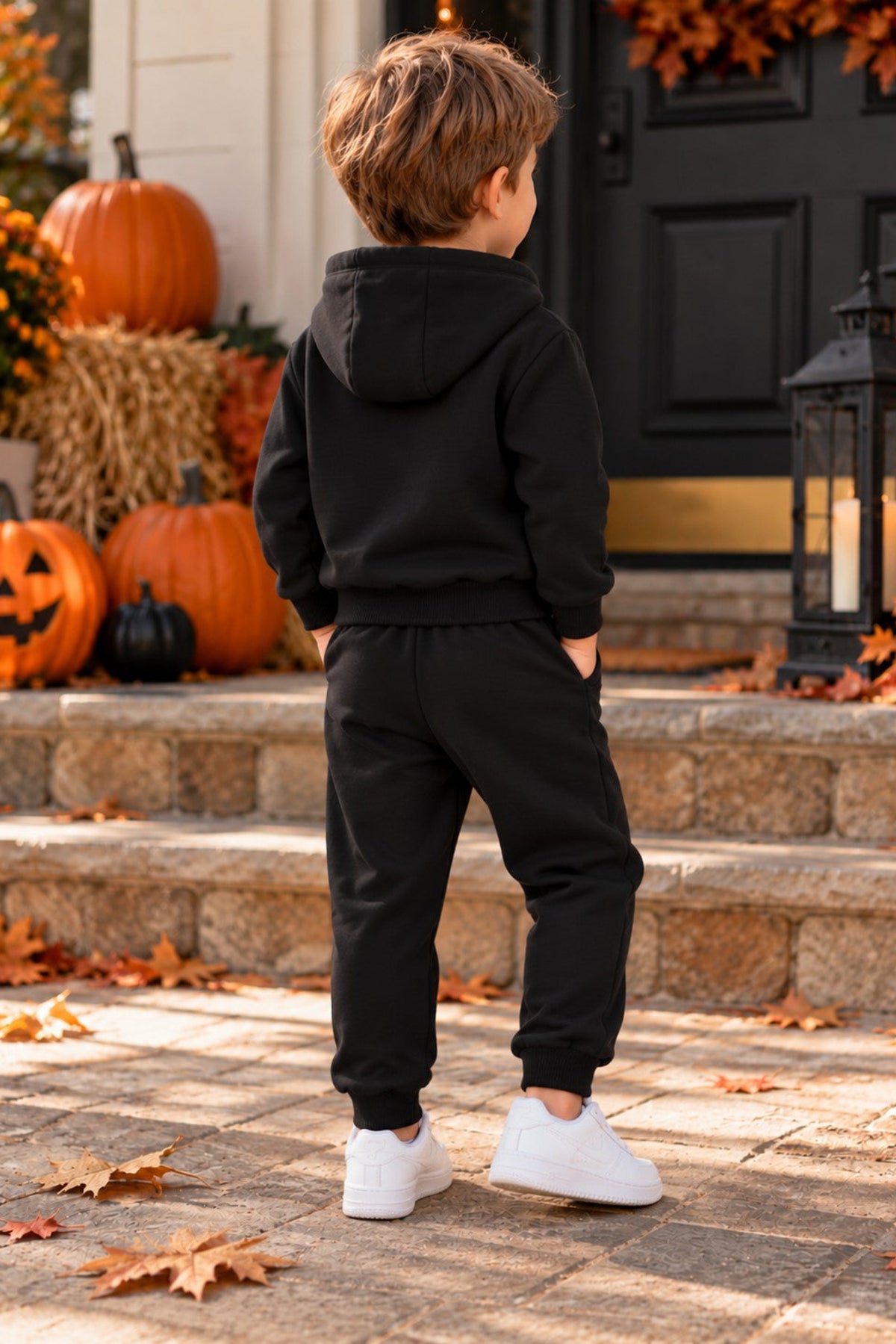 A young boy stands on stone steps in front of a decorated front door, wearing a black pullover hoodie and black jogger sweatpants with white sneakers. Pumpkins, autumn leaves, and a black lantern are visible in the background.