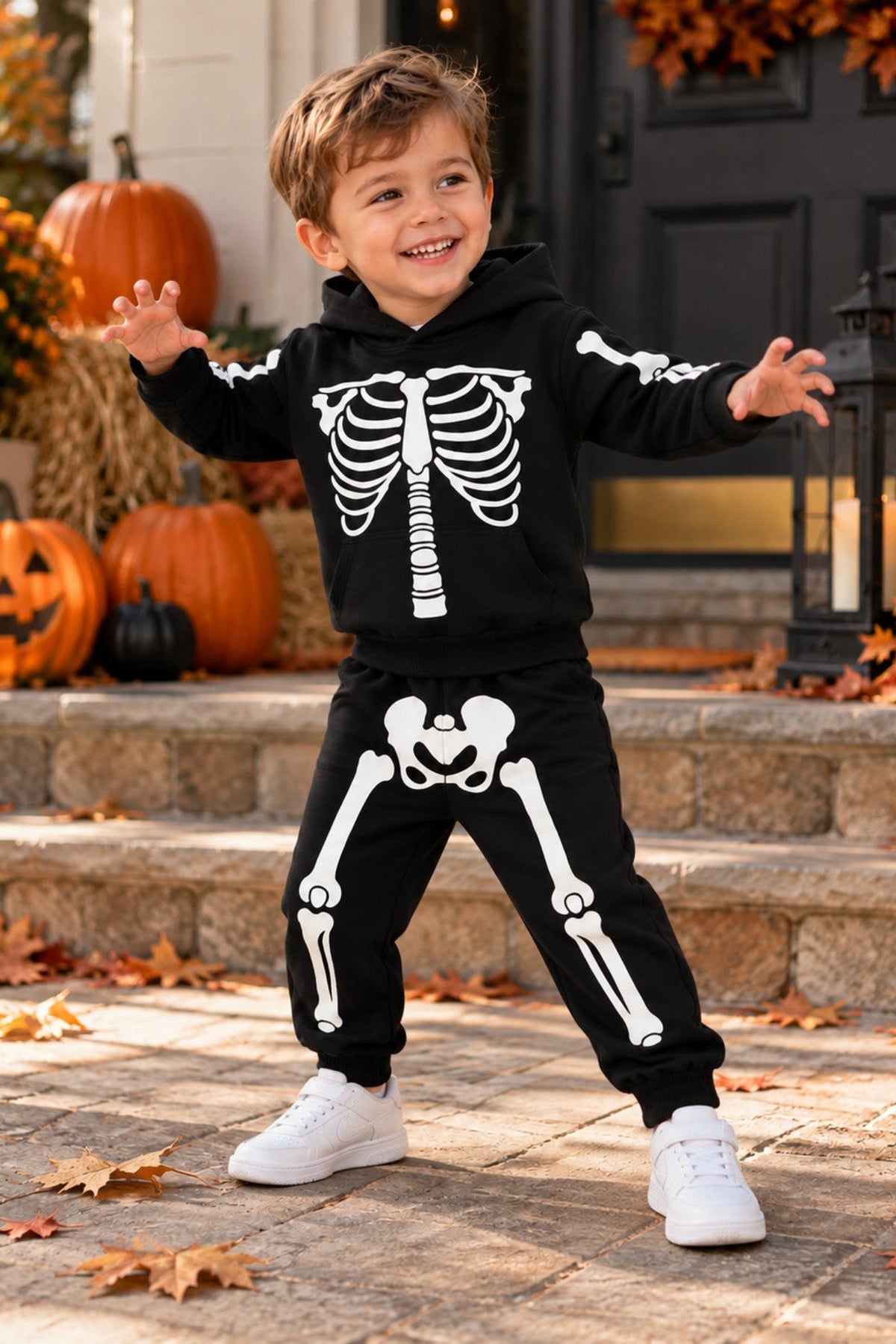 A young boy wearing a black and white skeleton print hoodie and sweatpants set, posing outdoors on stone steps decorated with pumpkins and autumn leaves.