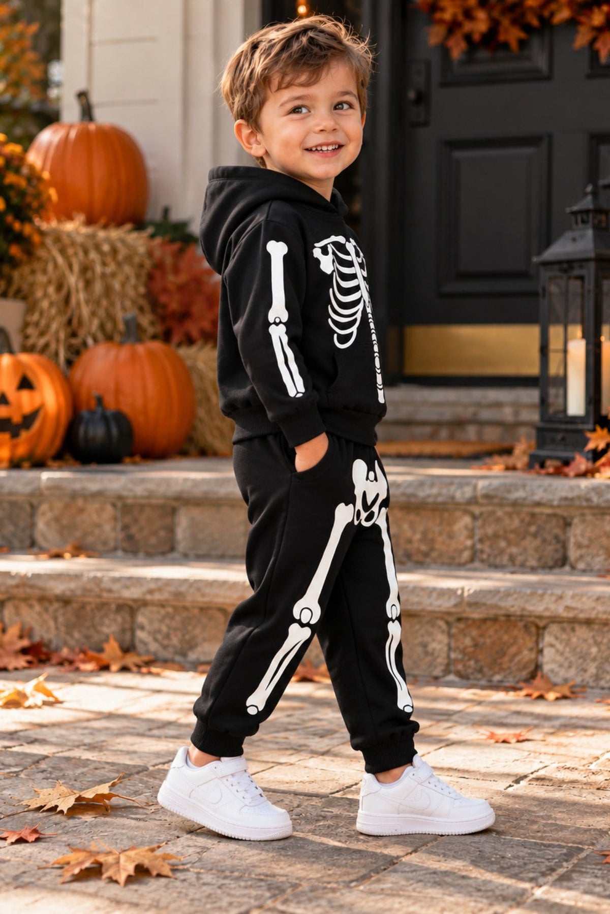 A young boy wearing a black skeleton-print hoodie and matching sweatpants stands on a stone porch decorated with pumpkins, autumn leaves, and a lantern.