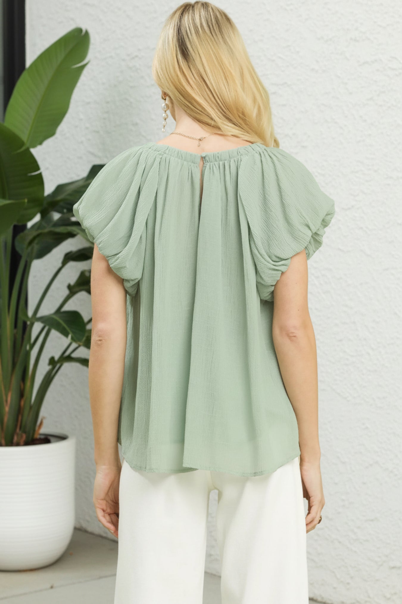 Woman wearing a light green blouse with ruffled sleeves against a white wall.