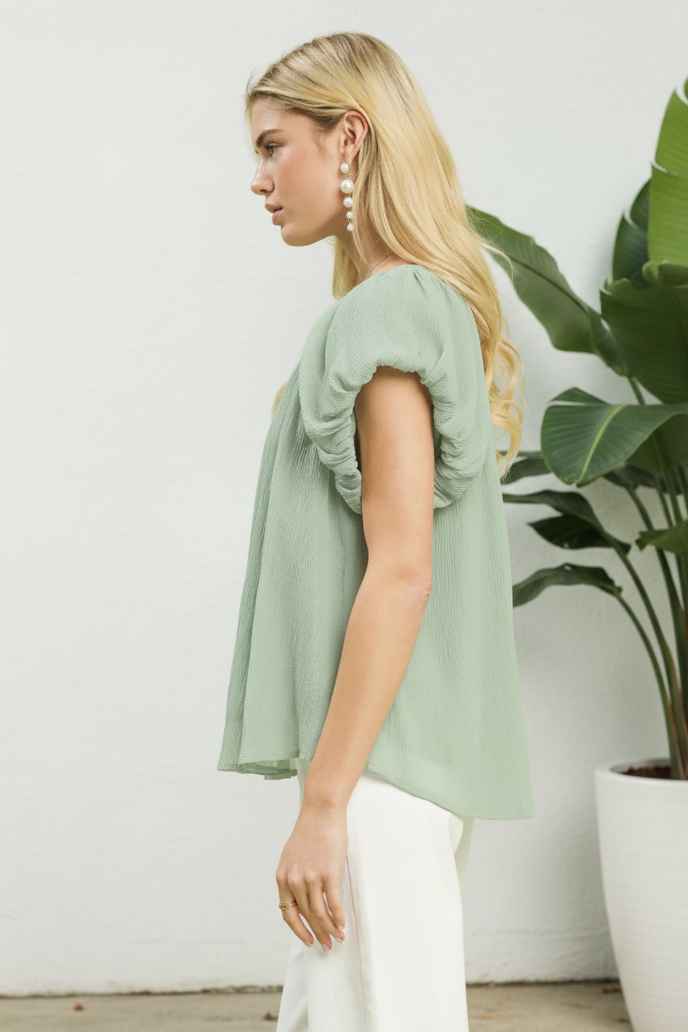 Woman wearing a light green blouse standing next to a potted plant against a white wall.