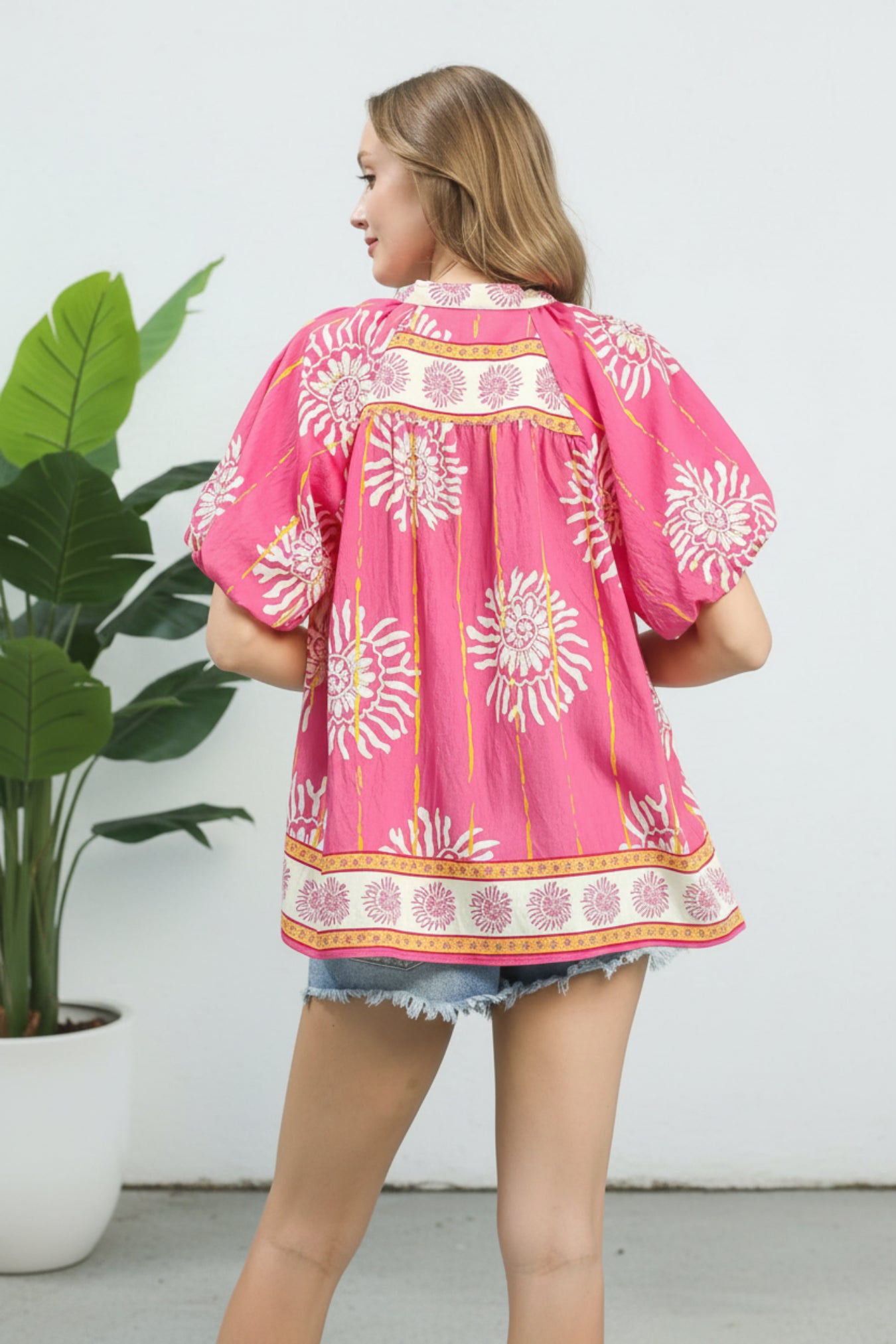 Woman wearing a pink floral blouse standing next to a potted plant indoors.