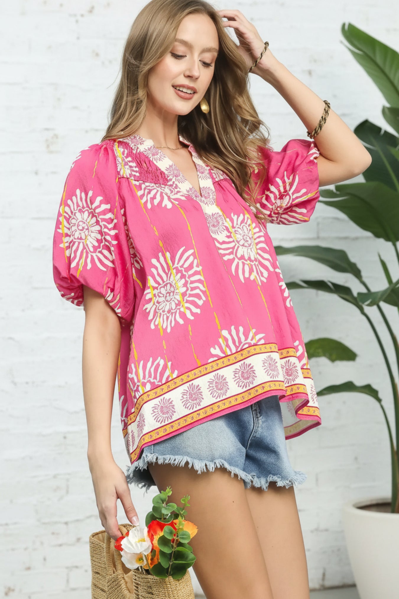 Woman wearing a pink floral blouse and denim shorts standing against a white brick wall with a plant in the background.