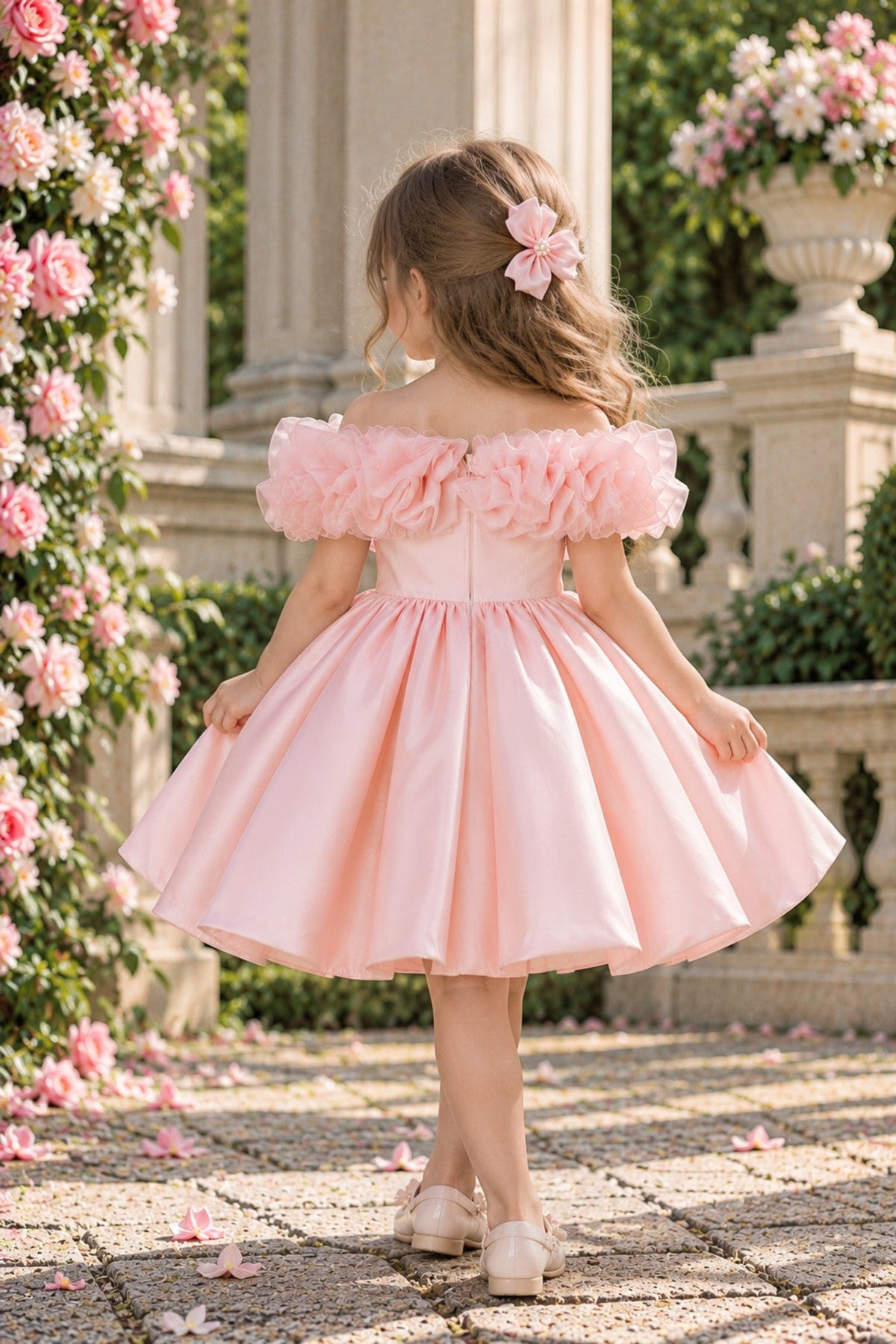 A young girl viewed from behind wearing a light pink off-the-shoulder flower girl dress with a ruffled bodice and a full skirt, standing in a garden courtyard with stone columns and pink rose vines.