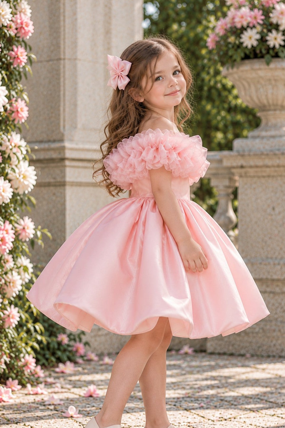 A young girl with long brown hair wearing a light pink off-the-shoulder ballgown and white ballet flats, standing in a garden courtyard with stone columns and pink flowers.