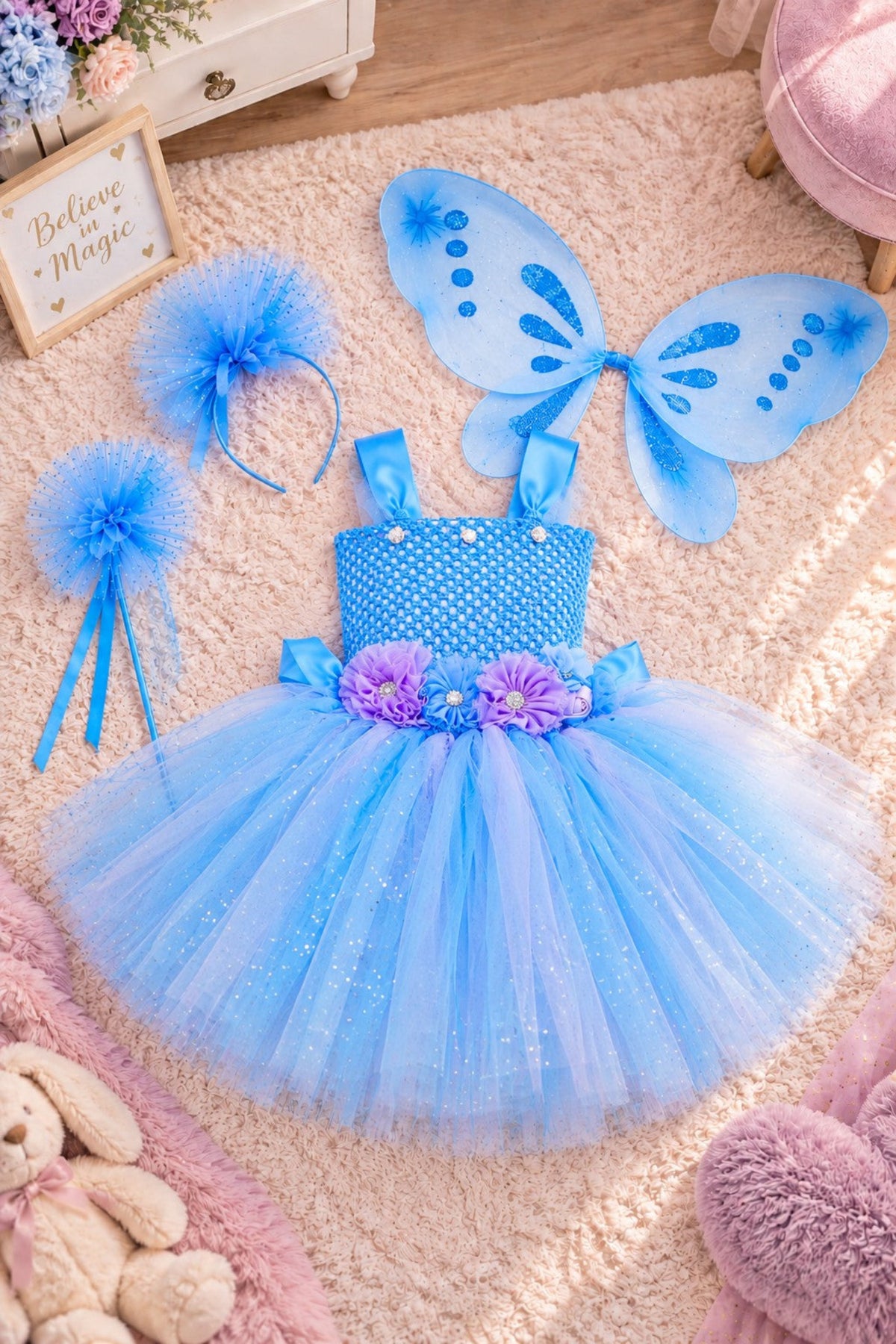 A children's fairy costume set laid out on a beige rug, including a light blue tutu dress with purple flower accents, matching blue wings, a blue headband, and blue pom-pom accessories.