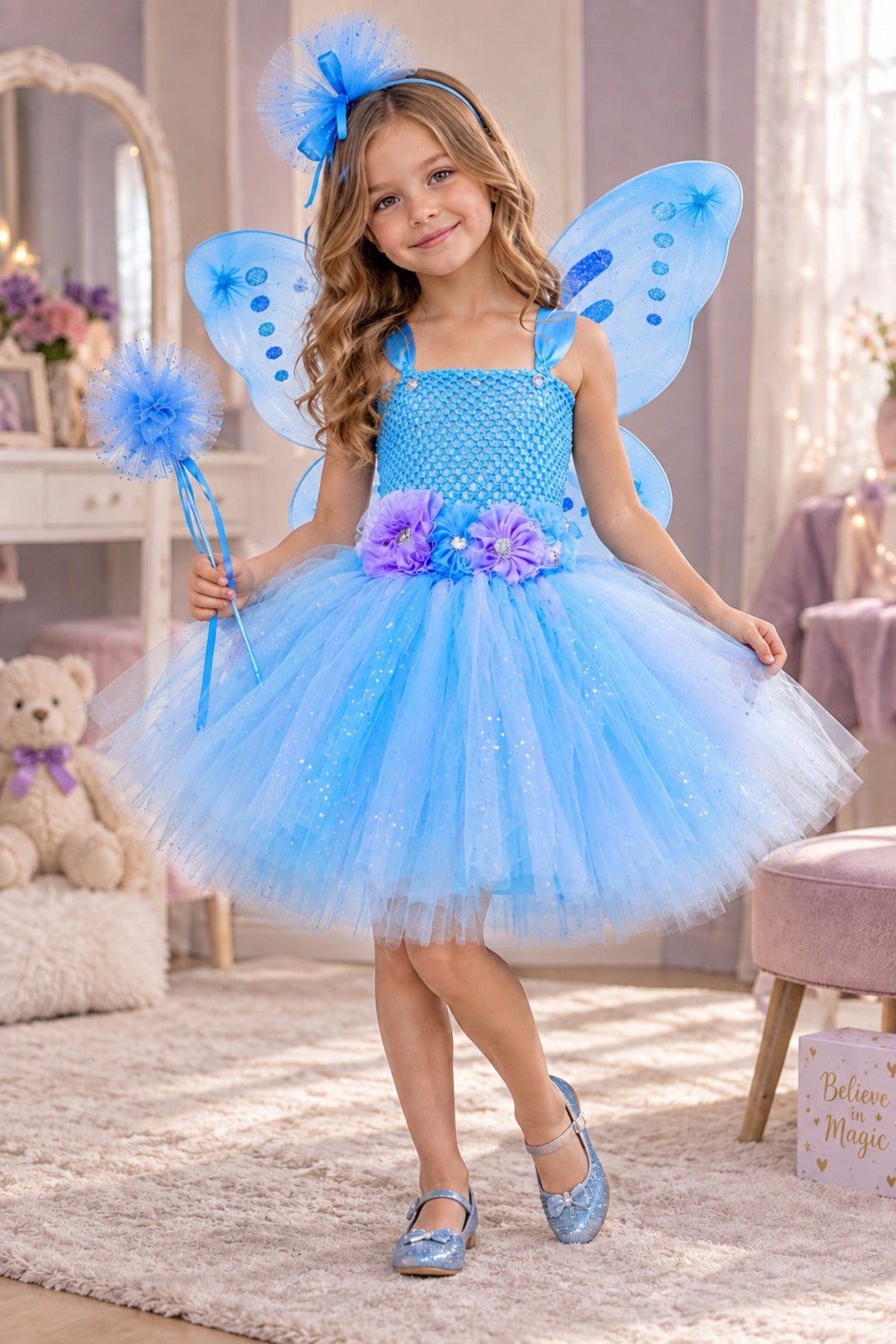 A young girl wearing a sparkly light blue fairy costume with matching blue wings, a blue bow headband, and holding a blue wand in a decorated children's room.