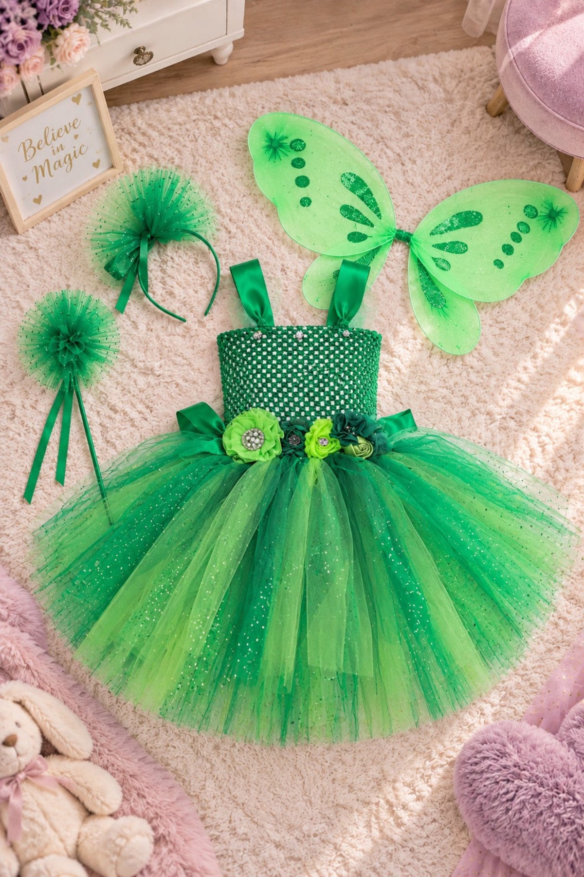A children's fairy costume set laid out on a soft pink rug, including a green tutu dress with a polka-dot bodice, matching green headband, fluffy green pom-poms, and green butterfly wings.