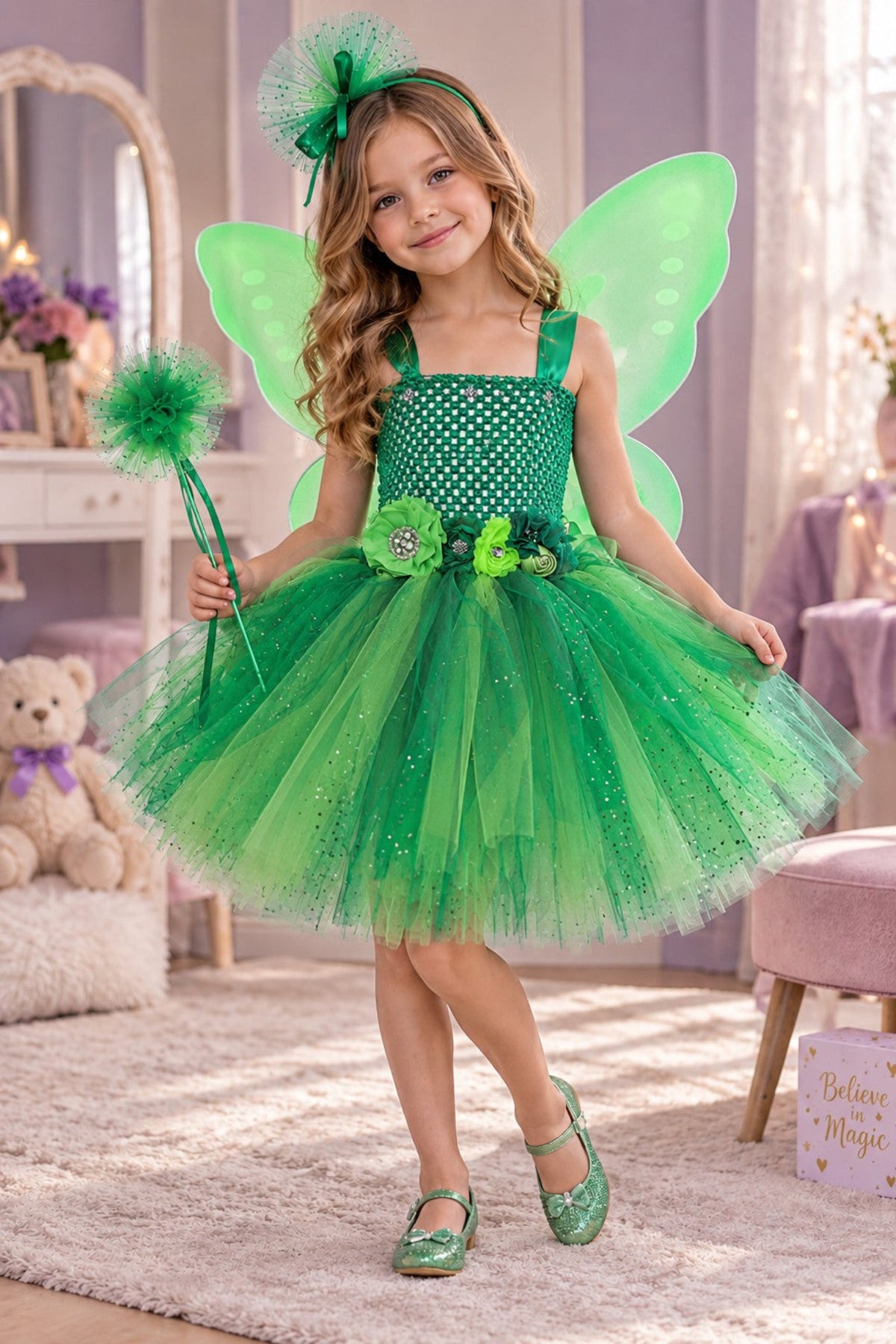 A young girl wearing a green fairy costume with a polka-dot bodice, tulle skirt, green wings, and a matching headband, standing in a decorated children's room and holding a wand.