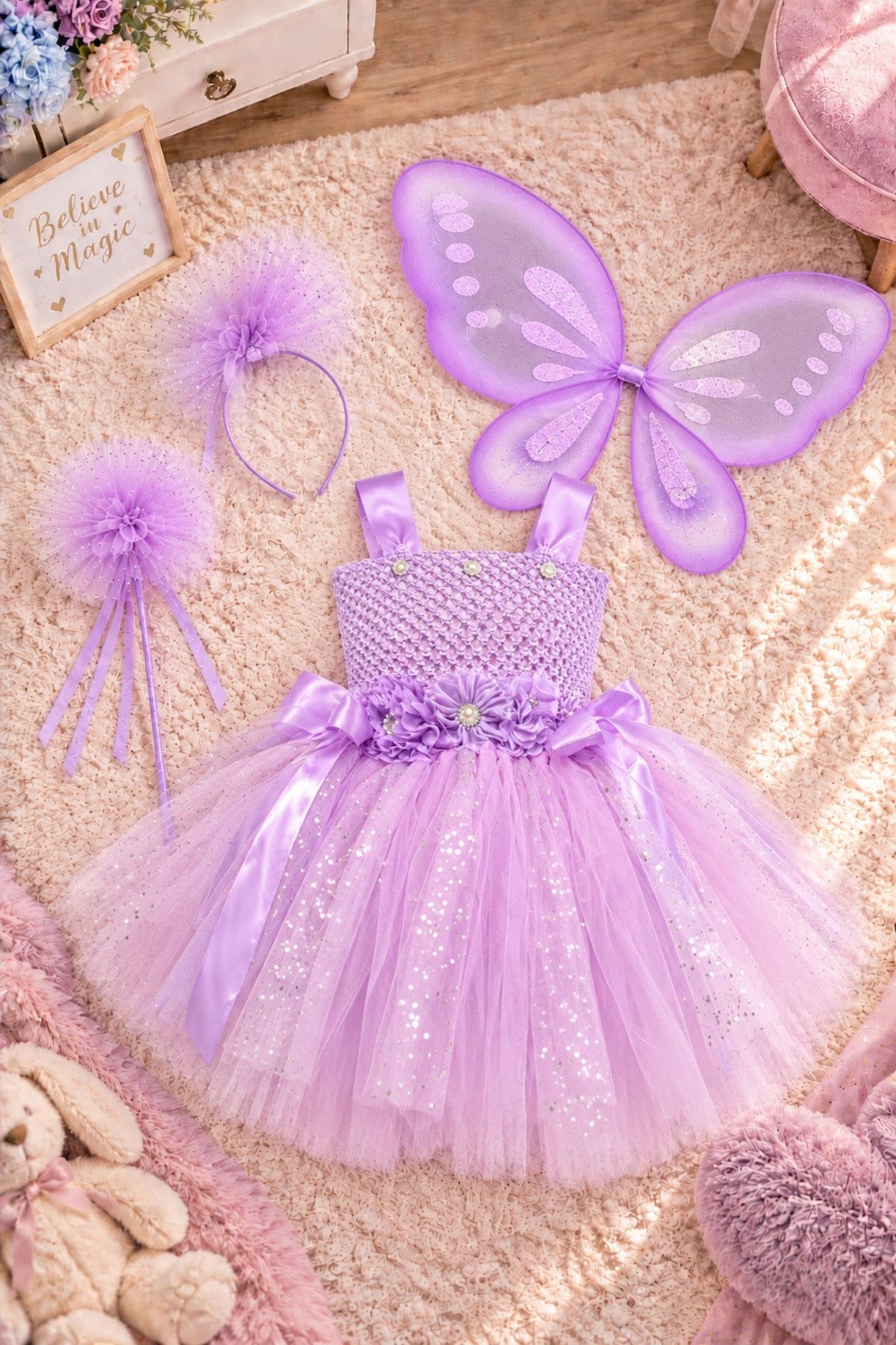 A children's fairy costume set laid out on a beige rug, including a lavender tulle dress with a sparkly bodice, matching butterfly wings, a purple pom-pom headband, and a purple wand with fluffy pom-poms.