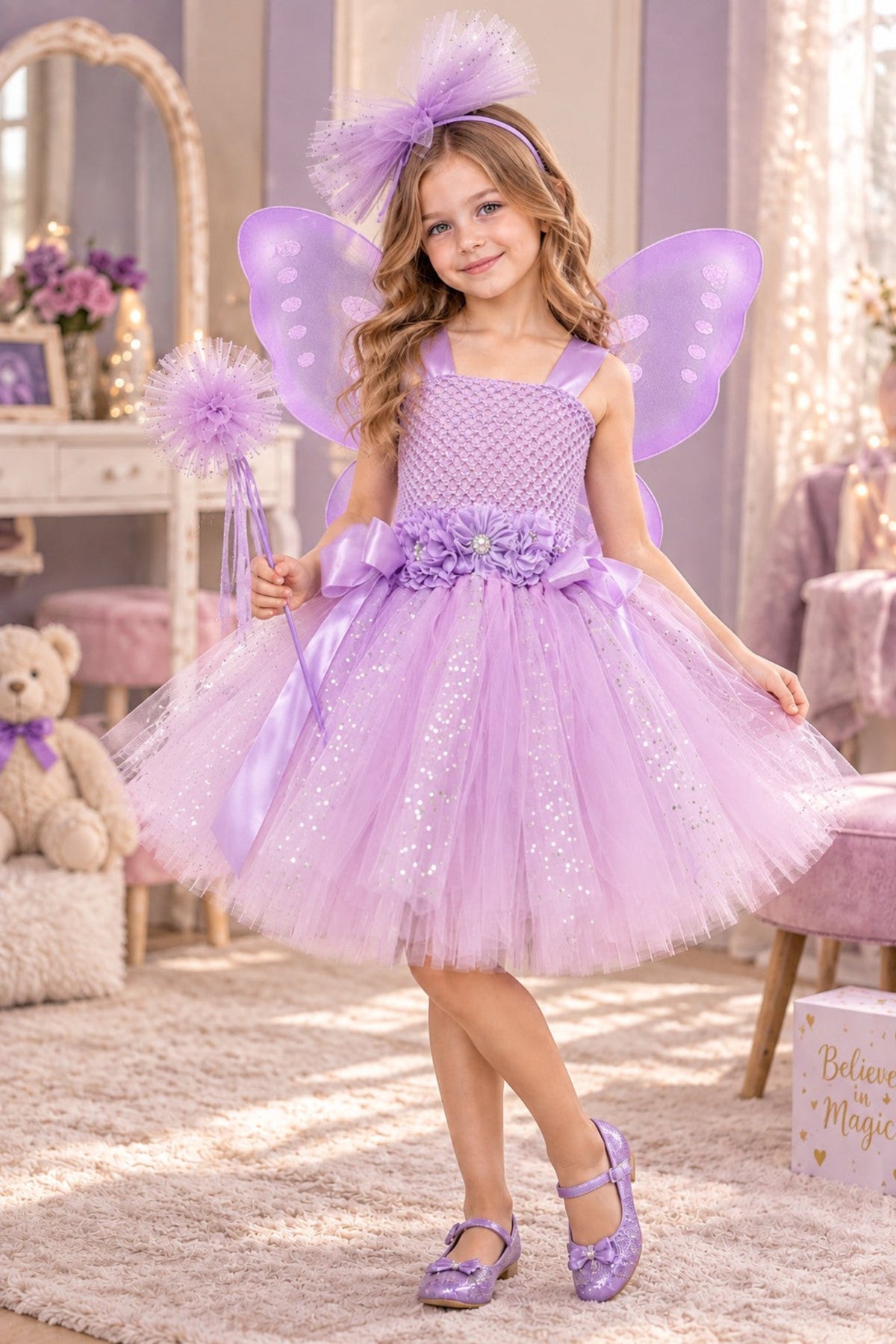 A young girl wearing a lavender fairy costume with a sparkly tulle dress, matching wings, a headband, and purple shoes, standing in a decorated room with a teddy bear and pink furniture.