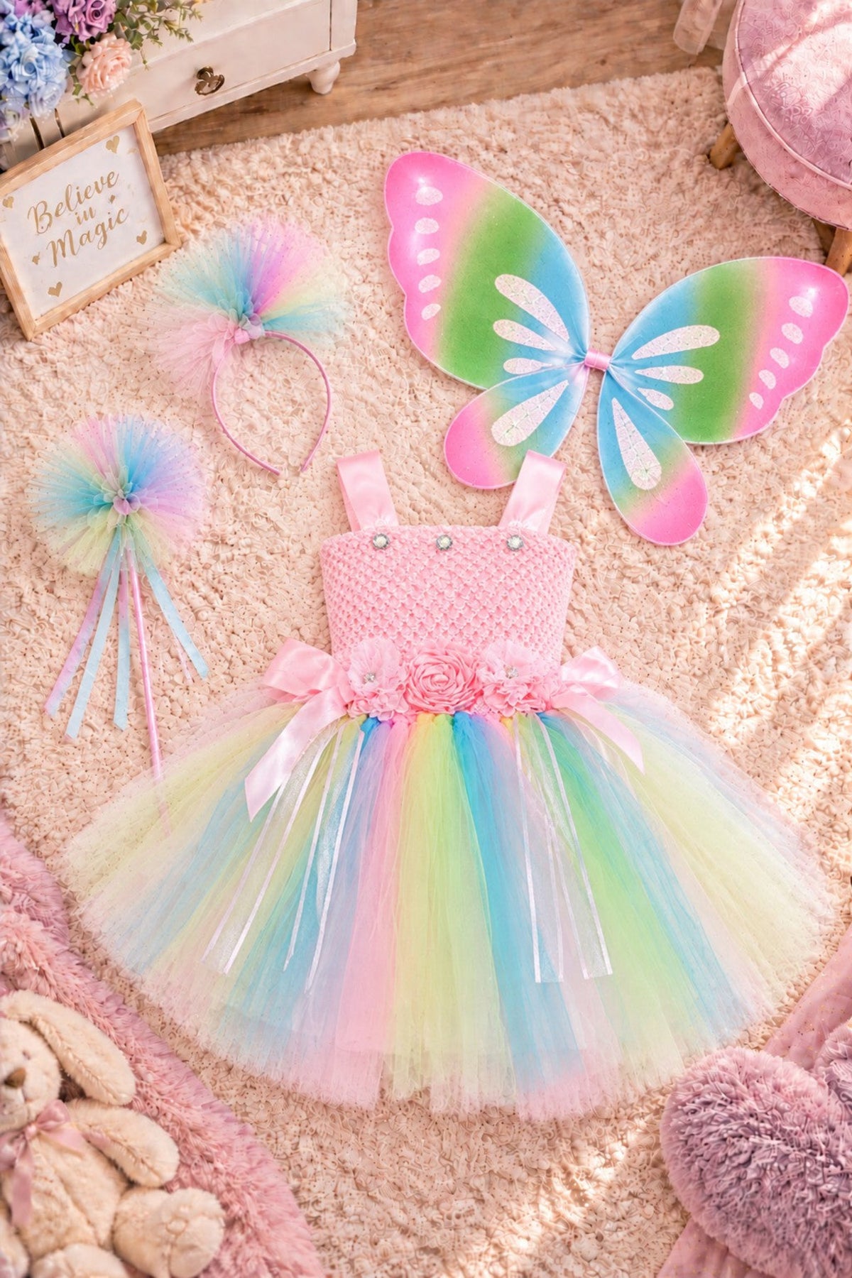 A children's pastel fairy costume set laid out on a soft pink rug, including a pink sleeveless tutu dress with a rainbow tulle skirt, a butterfly wing pair, a pink headband with a fluffy pastel pom, and a wand with matching pastel tulle.