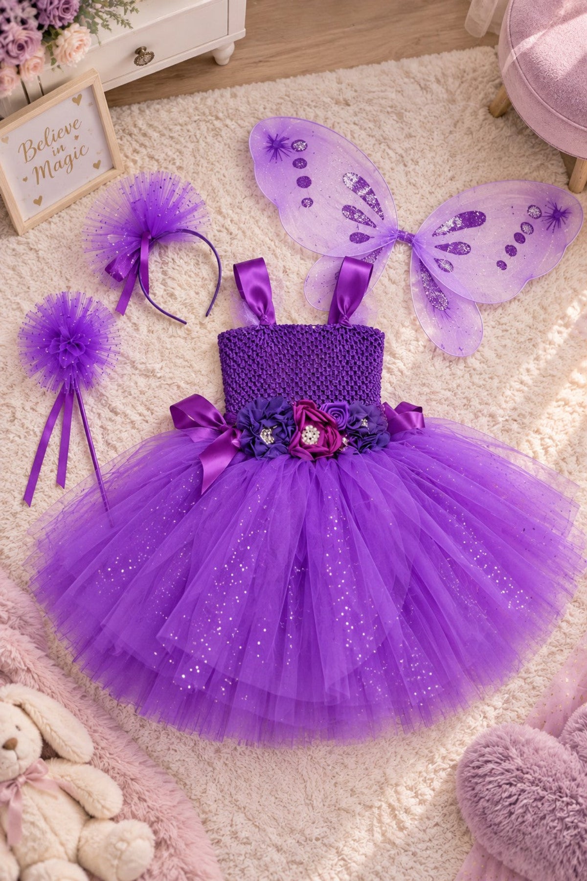 A purple toddler fairy costume set laid out on a cream rug, including a sparkly tulle tutu dress with a purple bodice, matching purple wings, a tulle headband, a pom-pom headband, and a purple bow headband.
