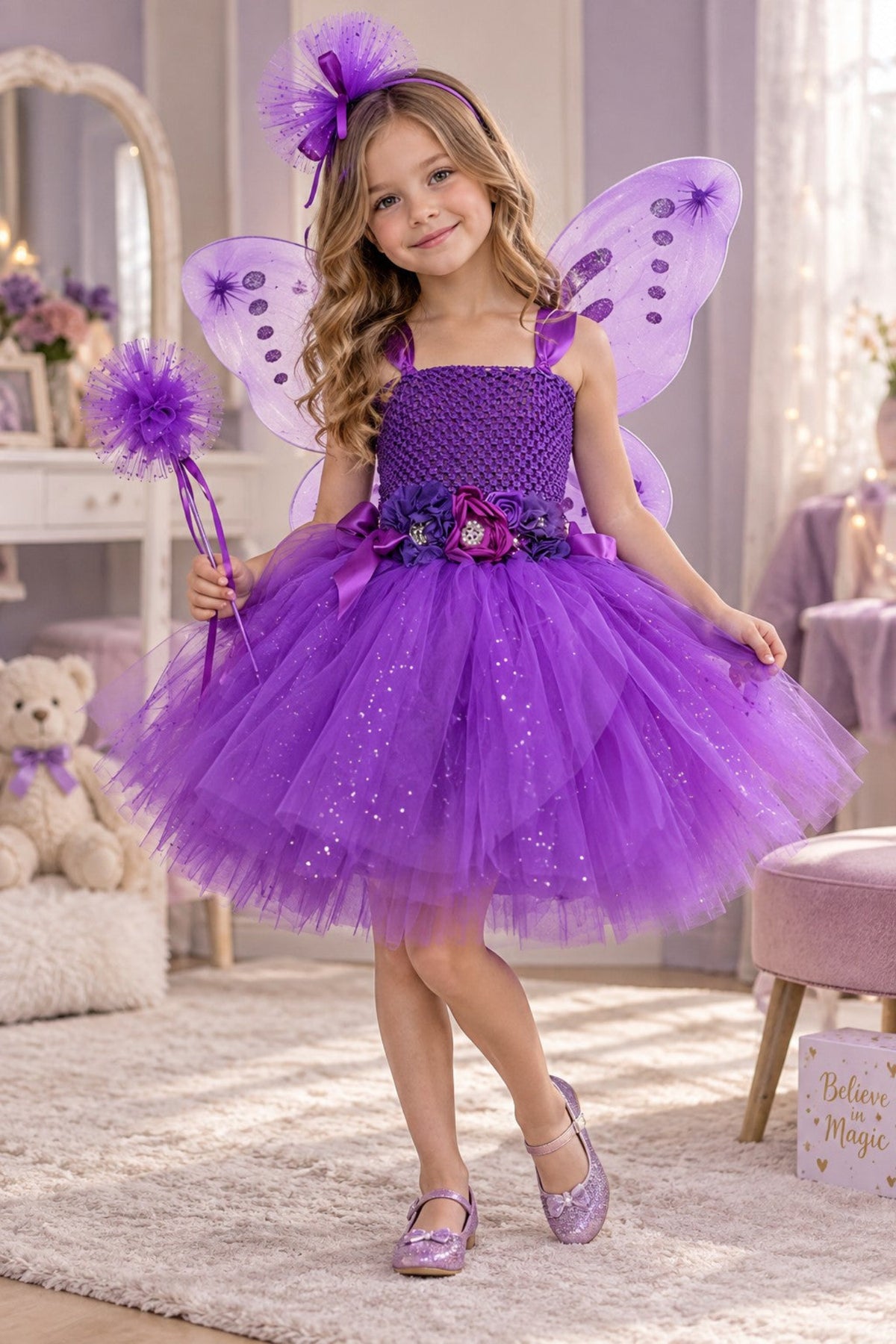 A young girl wearing a sparkly purple fairy costume with matching purple wings, a headband, and a wand, posing in a decorated children's room.