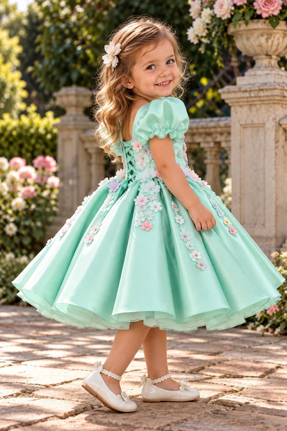 A young girl with blonde hair wearing a mint green party dress with 3D floral appliqués and white ballet flats, standing in a garden courtyard.