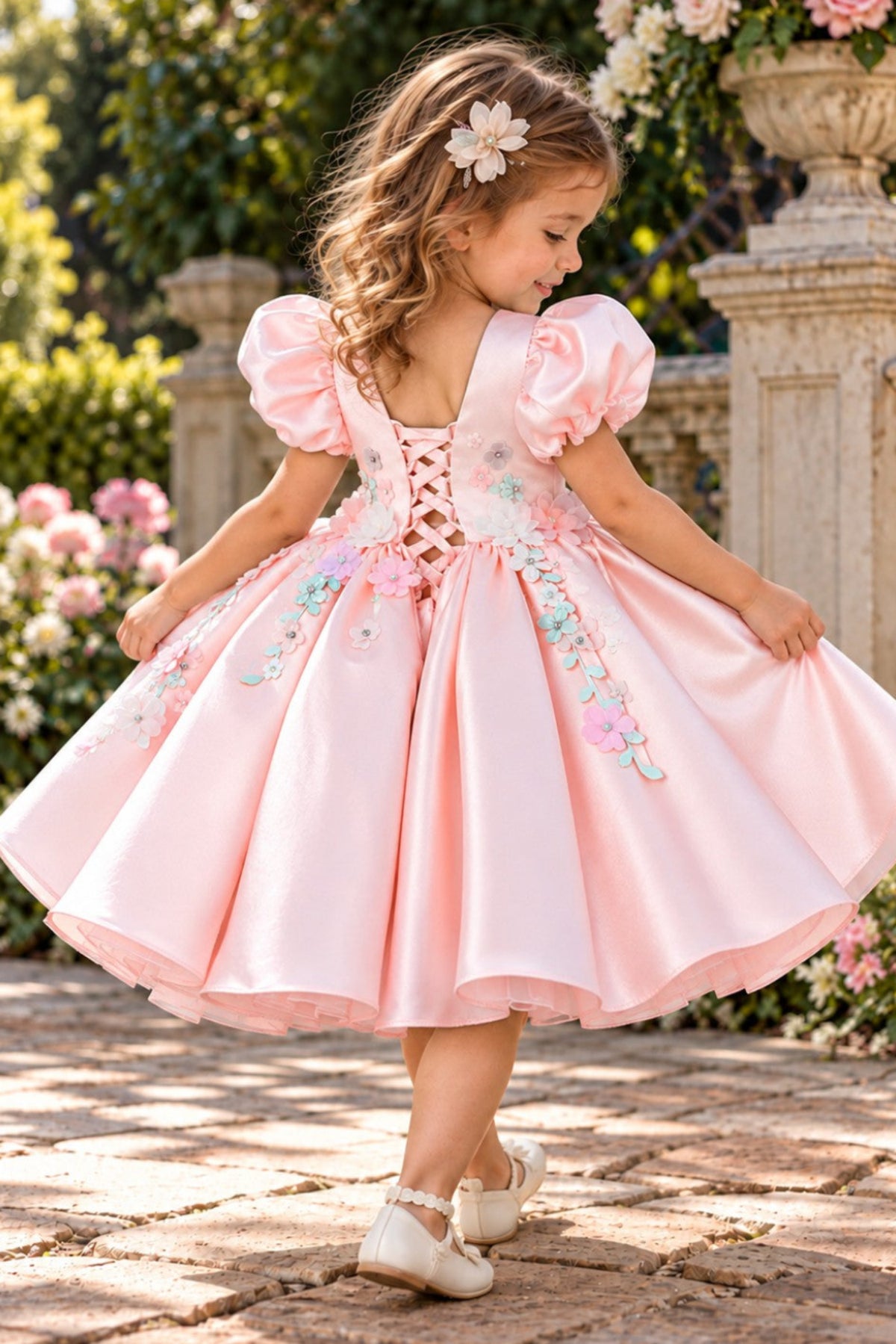 A young girl wearing a light pink satin ballgown with puff sleeves, a lace-up back, and floral embroidery, standing in a garden courtyard.