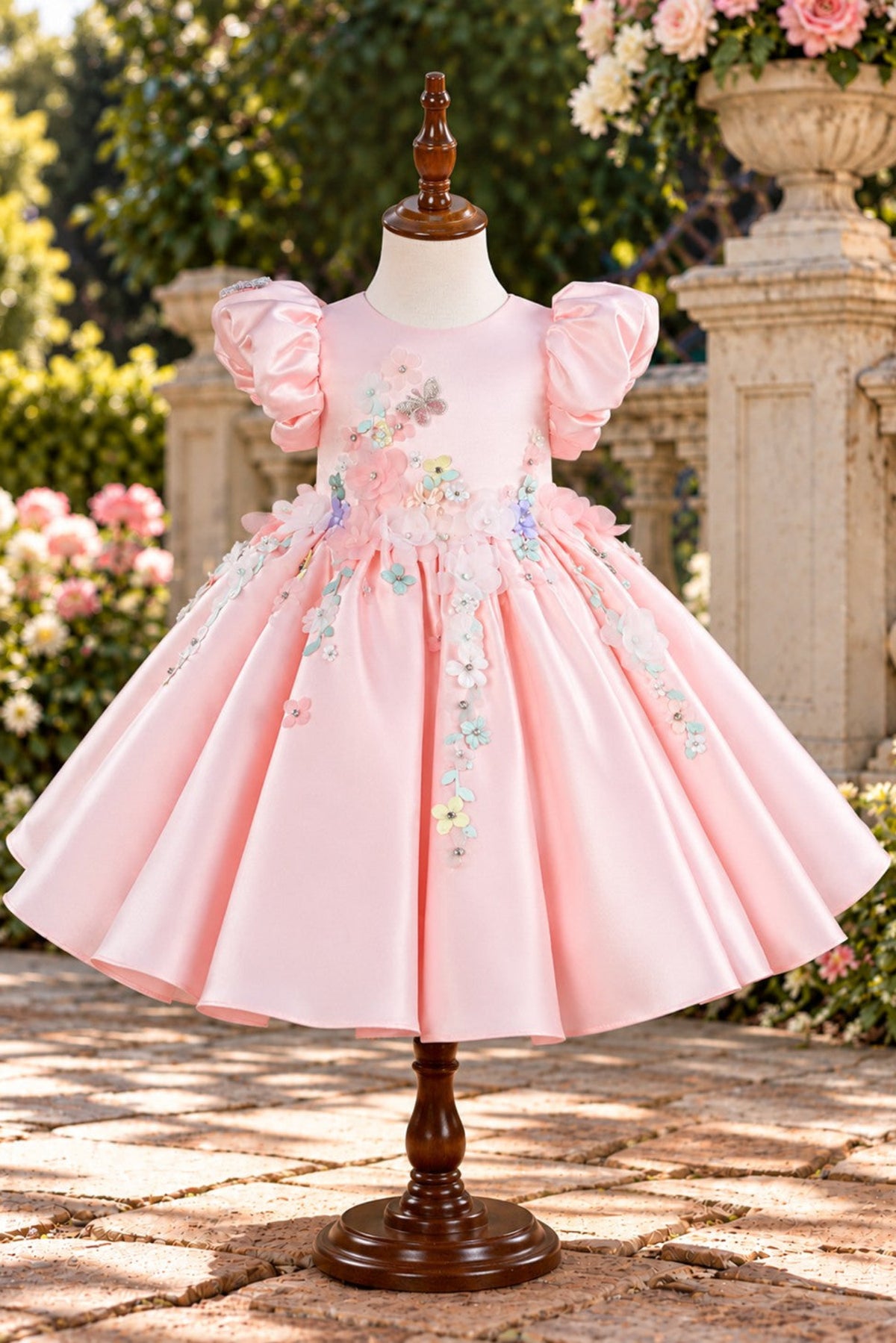 A pink toddler girl's dress displayed on a mannequin outdoors. The dress has puffed short sleeves, a round neckline, a full skirt, and delicate floral embroidery with butterfly details on the bodice and upper skirt.