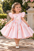 A young girl with curly brown hair wearing a pink satin dress with puff sleeves and colorful floral embroidery, standing on a stone path in a garden.