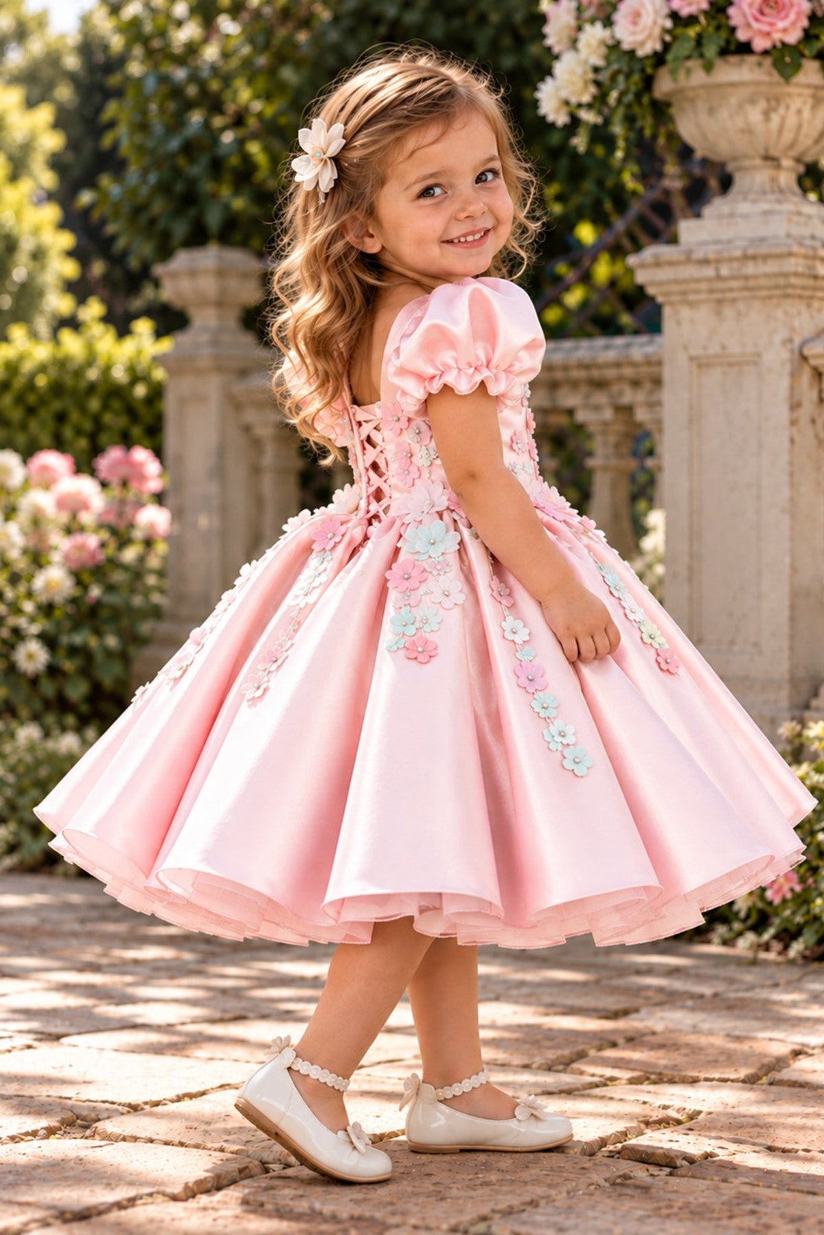 A young girl with blonde hair wearing a pink formal party dress with floral embroidery and puff sleeves, standing in a garden courtyard.