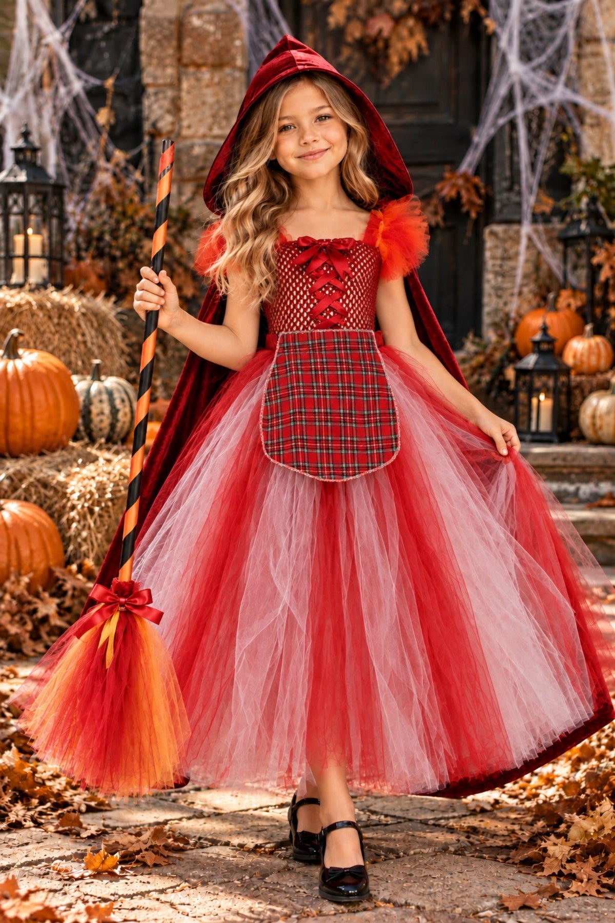 A young girl wearing a Red Riding Hood costume stands in an autumn-themed outdoor setting. The costume includes a red hood, a red and black plaid bodice with faux lace-up details, a red tulle tutu skirt, and red feather accents on the shoulders.