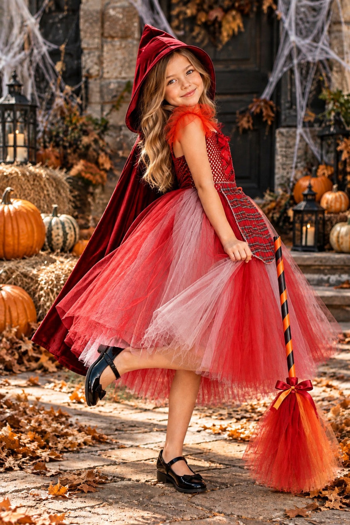 A young girl modeling a red Little Red Riding Hood costume in an outdoor autumn setting with pumpkins, hay bales, and cobwebs.