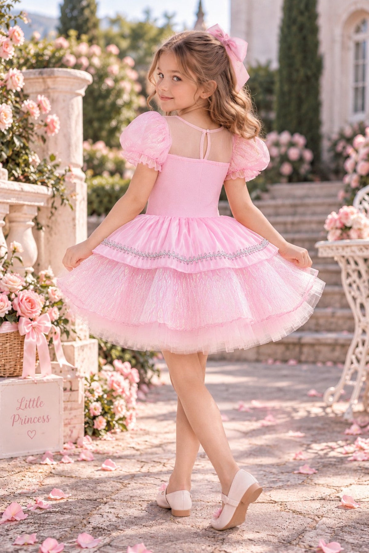 A young girl wearing a pink tiered tulle dress with puff sleeves and a matching pink bow in her hair, posing outdoors in a garden setting with stone steps and pink rose decorations.