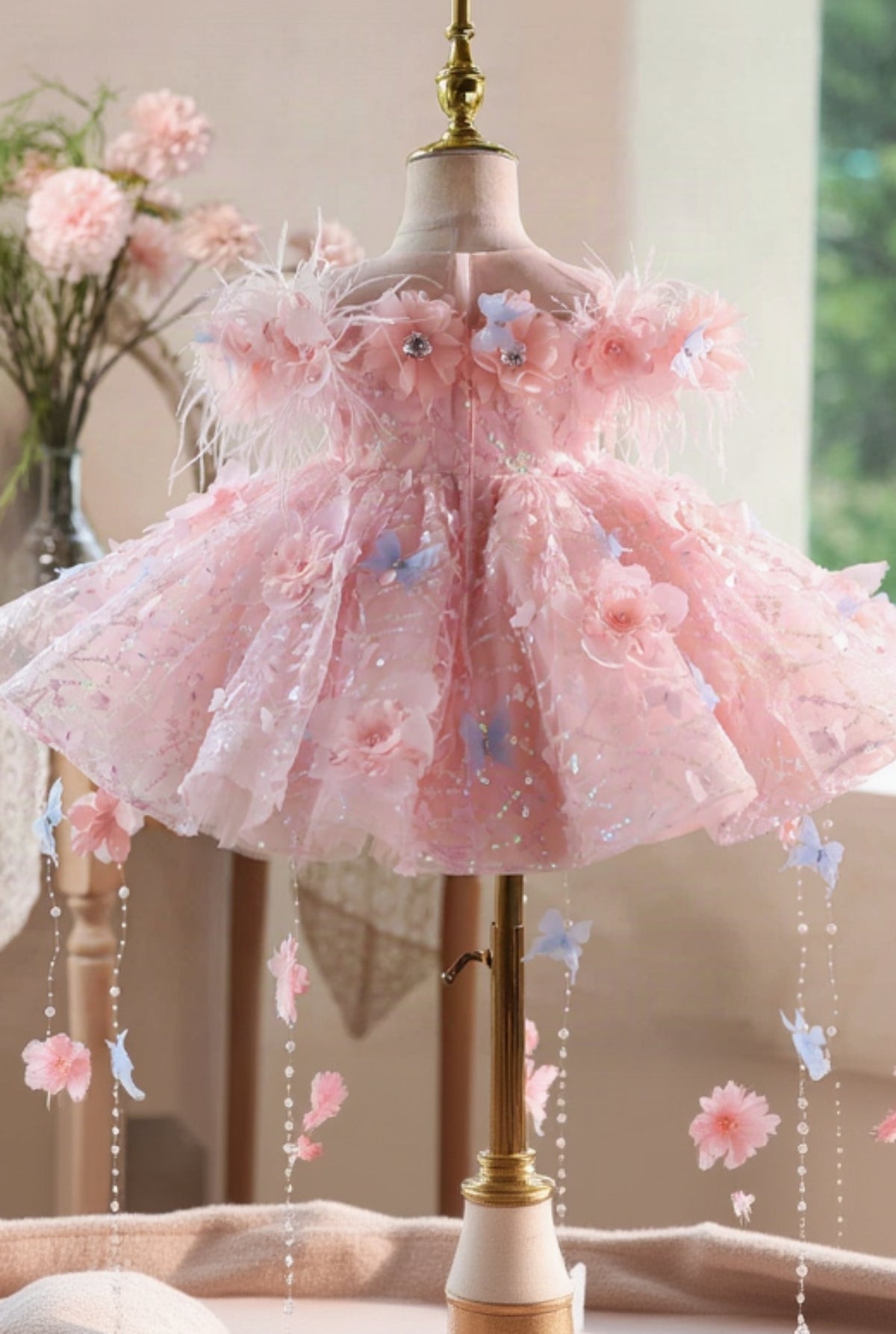 A pink girls' party dress displayed on a mannequin dress form. The dress features a sheer tulle bodice with 3D floral appliqués, feathered shoulder details, and a voluminous layered tulle skirt decorated with glitter, flowers, and hanging blue and pink butterfly charms.