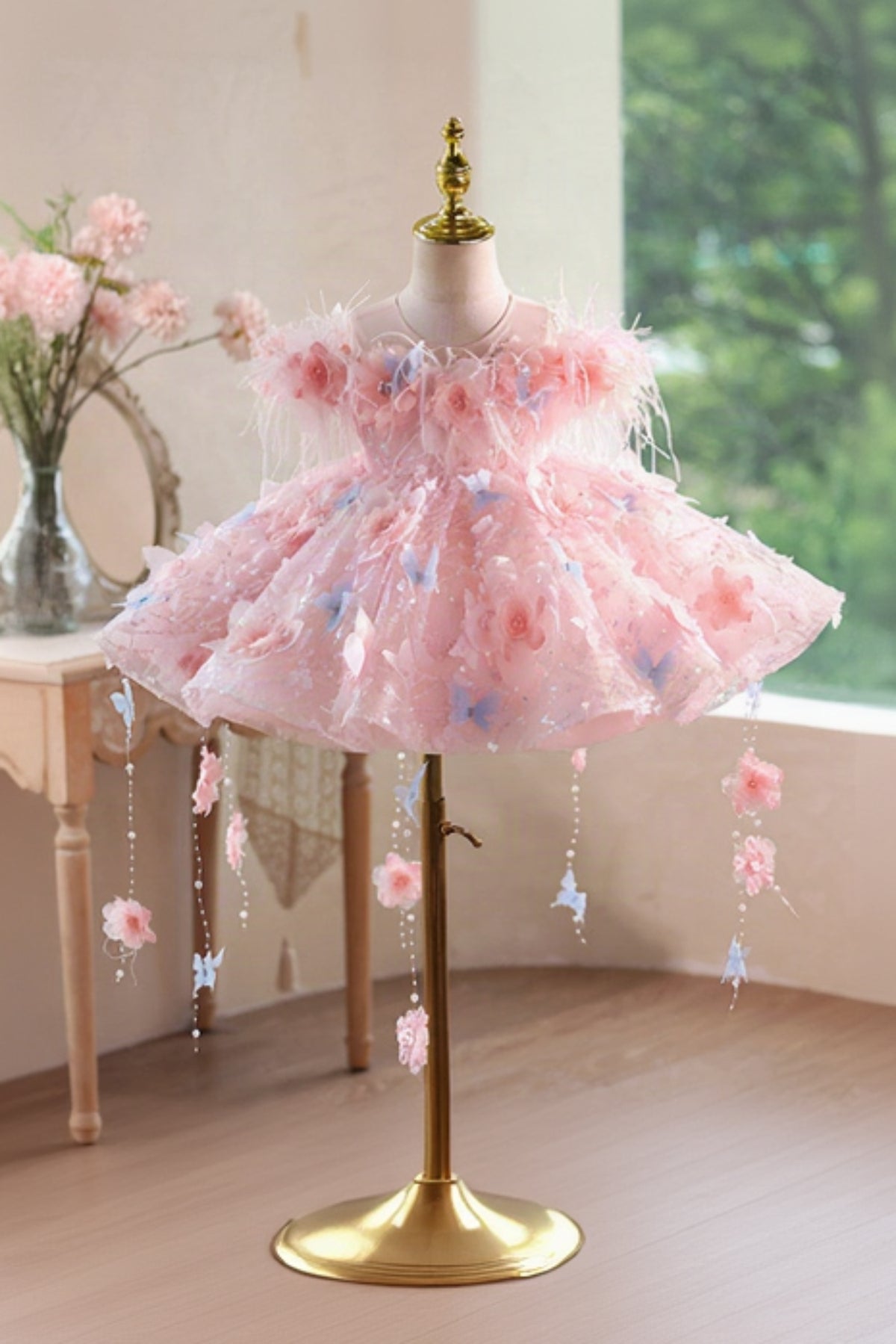 Pink children's dress with floral and butterfly details on a mannequin against a window backdrop.