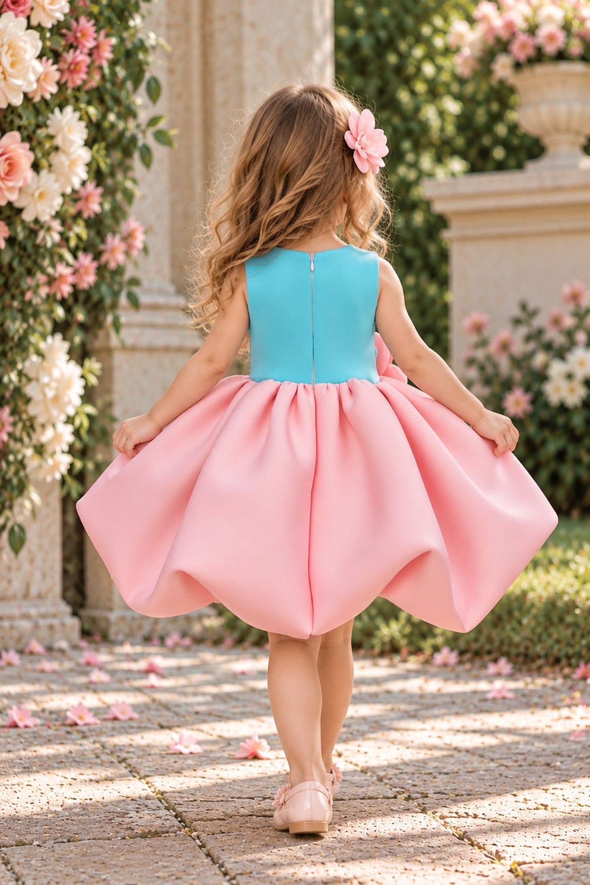 A young girl viewed from behind wearing a sleeveless two-tone dress with a turquoise blue bodice and a voluminous pink skirt, standing on a stone path in a garden with flowers and columns in the background.
