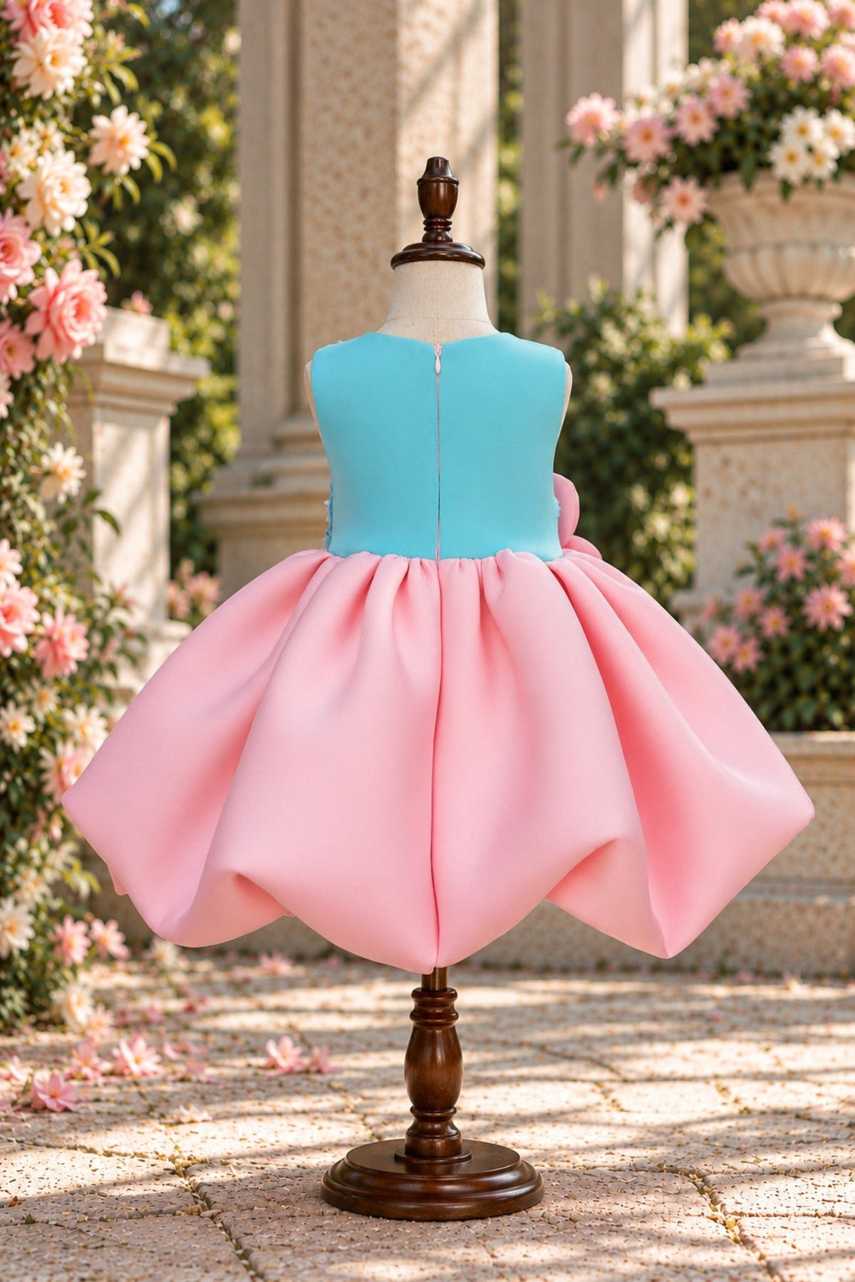 A two-piece girls' dress displayed on a mannequin outdoors. The dress has a bright turquoise blue sleeveless top with a center zipper and a voluminous light pink skirt with a matching pink bow at the waist.
