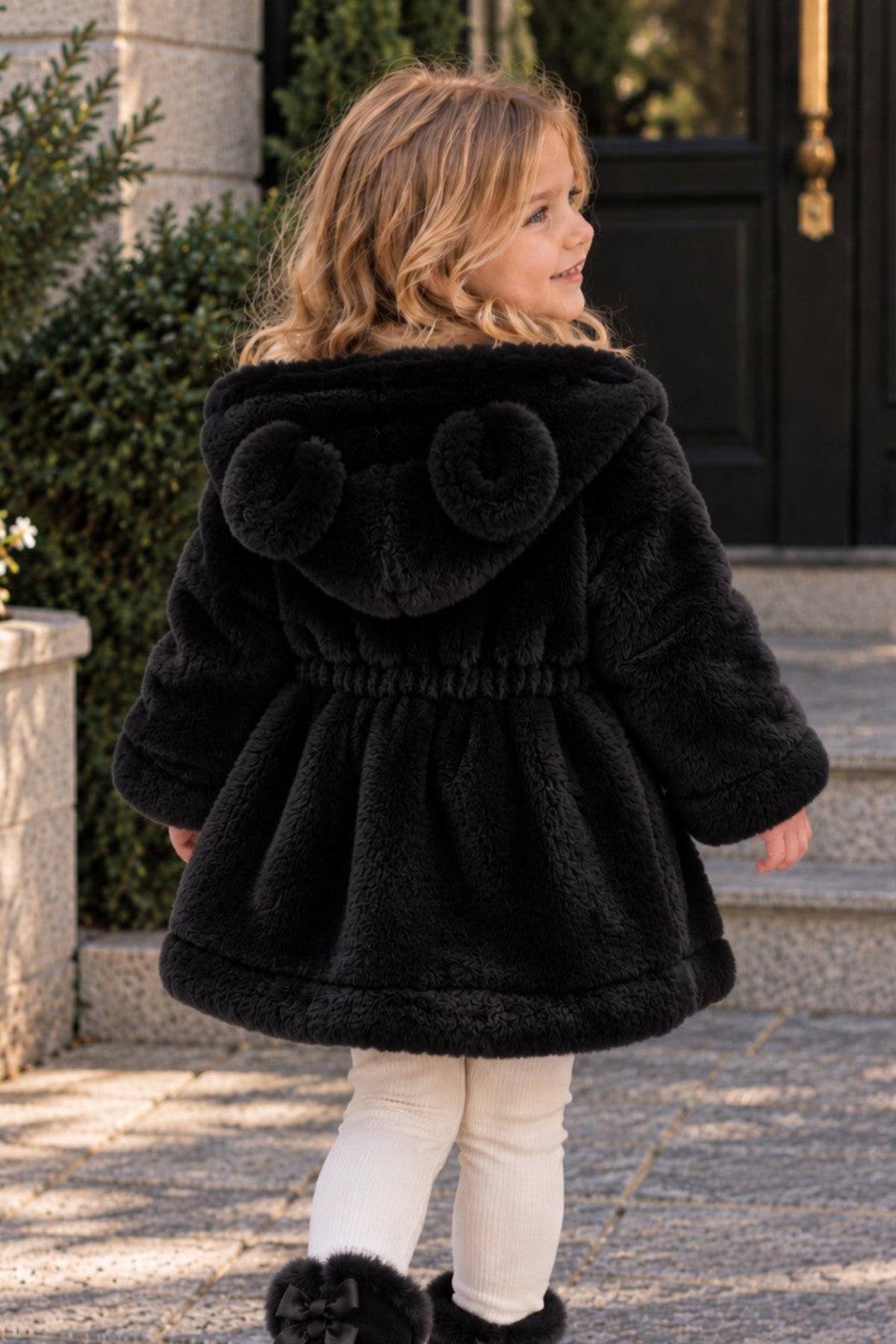 A young girl wearing a black faux fur hooded coat with bear ears on the hood, white leggings, and black boots, walking on a stone path outdoors.