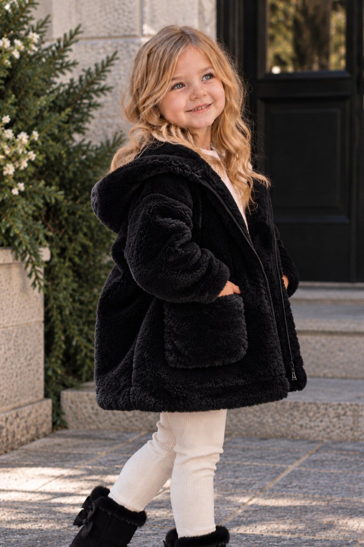 A young girl wearing a black fuzzy hooded jacket, white leggings, and black ankle boots with bows, standing on stone steps outdoors.