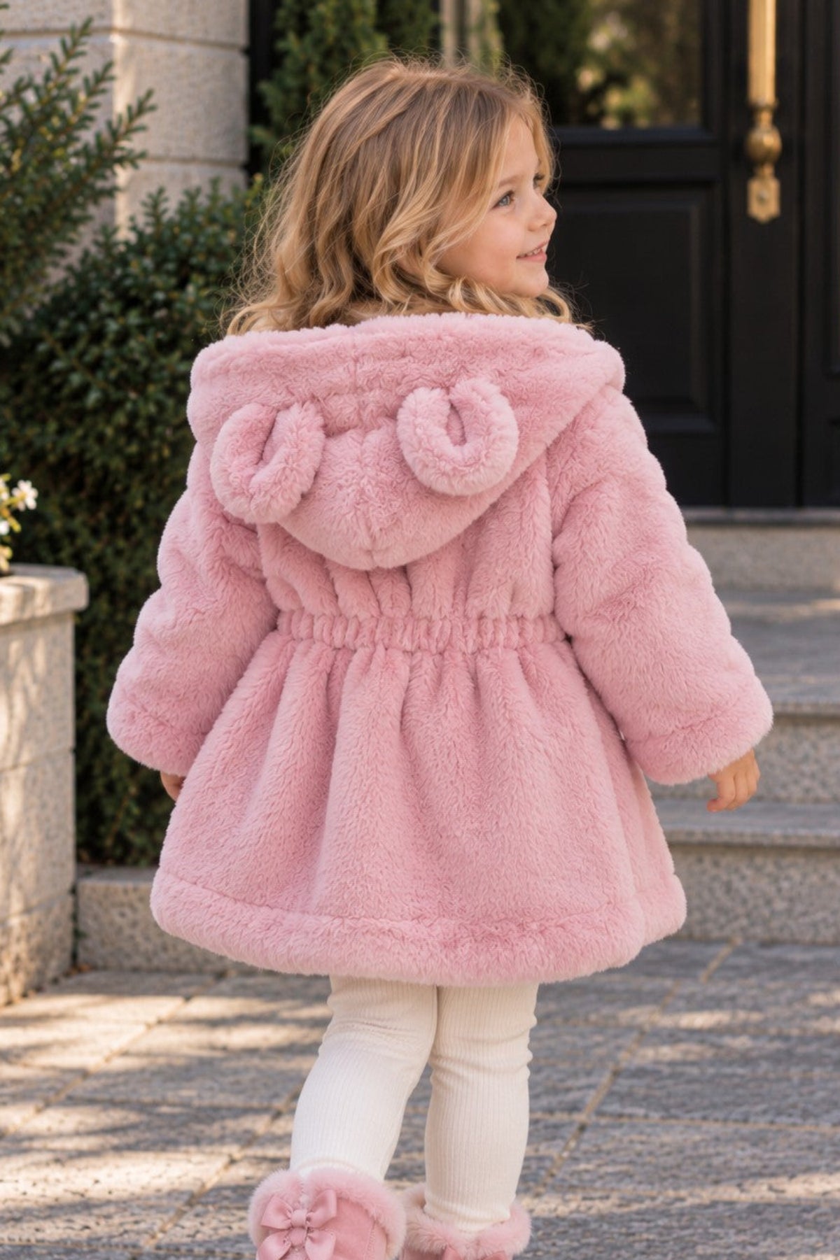 A young girl viewed from behind wearing a fluffy light pink hooded coat with large pom-poms on the hood, white leggings, and pink fuzzy boots with bows, standing on a stone walkway in front of a building.