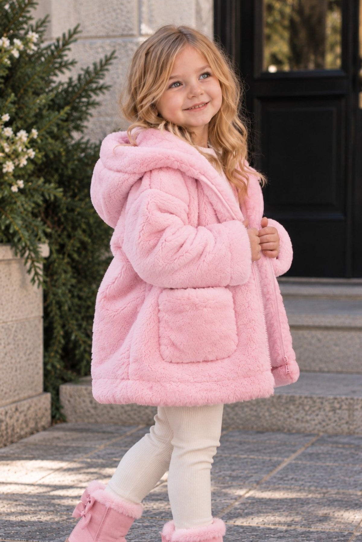 A young girl wearing a fluffy light pink hooded jacket, cream leggings, and matching pink fuzzy boots with bows, standing outdoors on stone steps in front of a building.